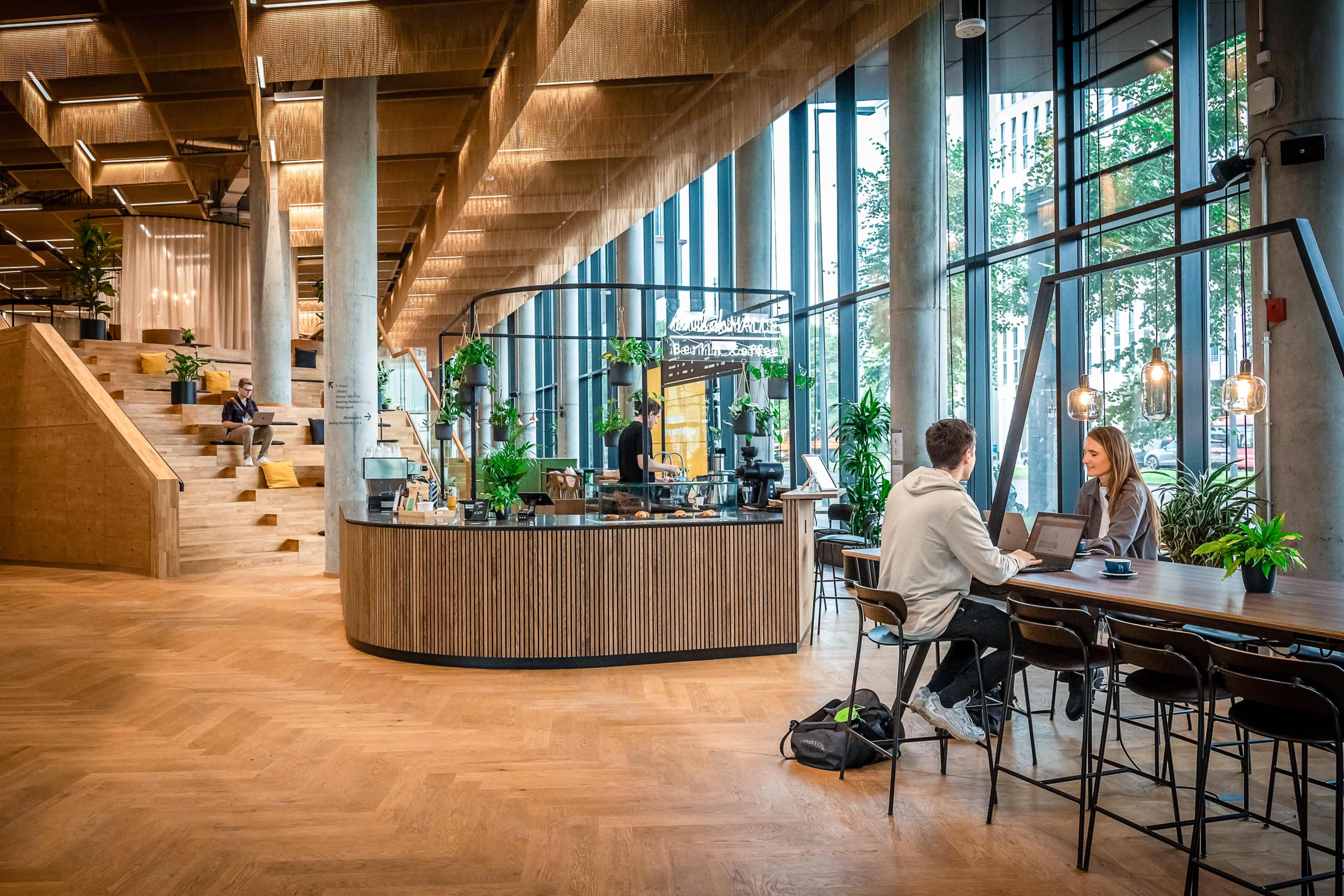 The image shows a modern interior space featuring a coffee bar with a curved counter, surrounded by wooden furnishings and greenery, along with people working at tables and seated in a lounge area.