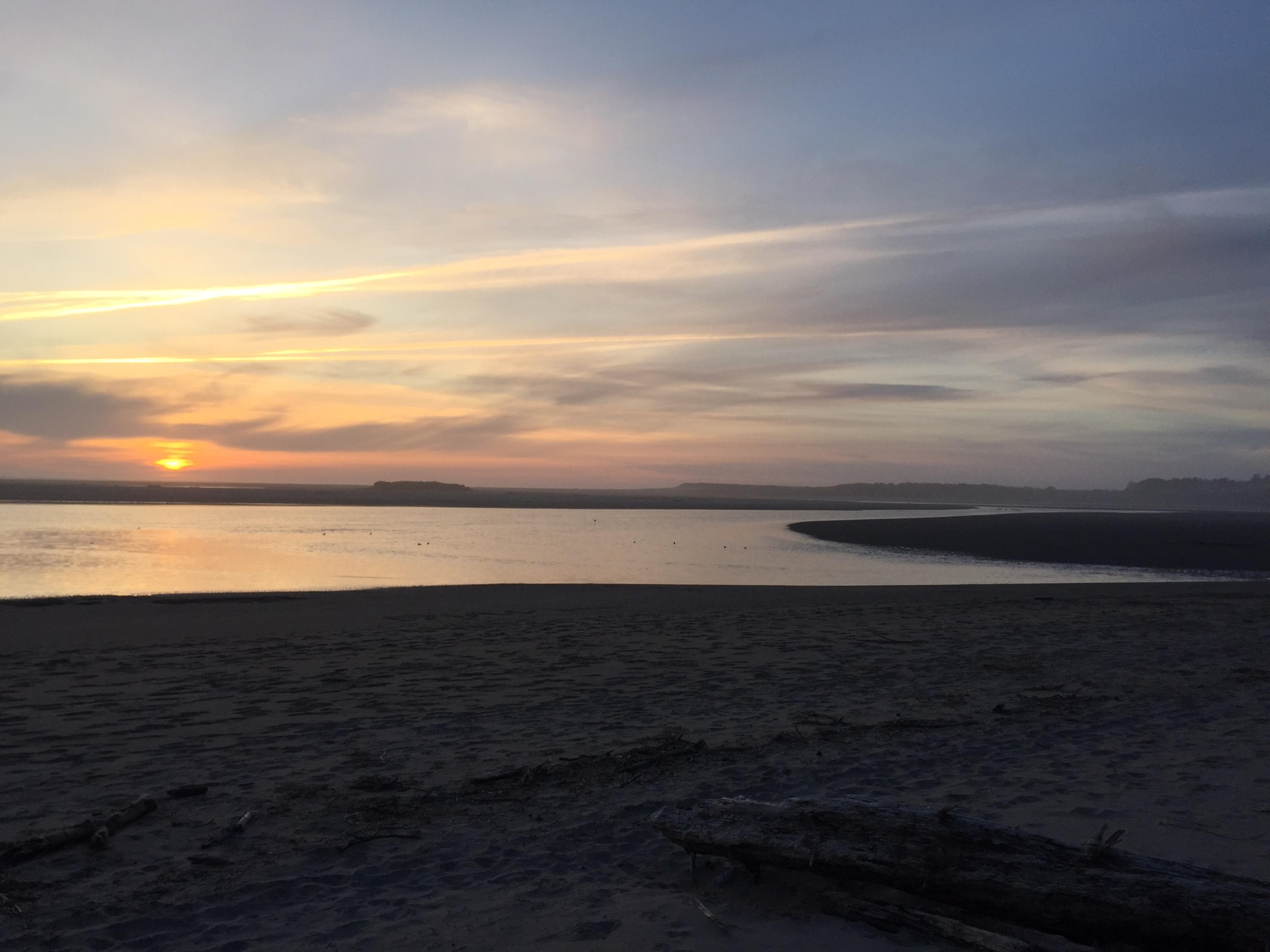 A tranquil beach at sunset, with a curve in the water and a silhouette of distant land on the horizon.