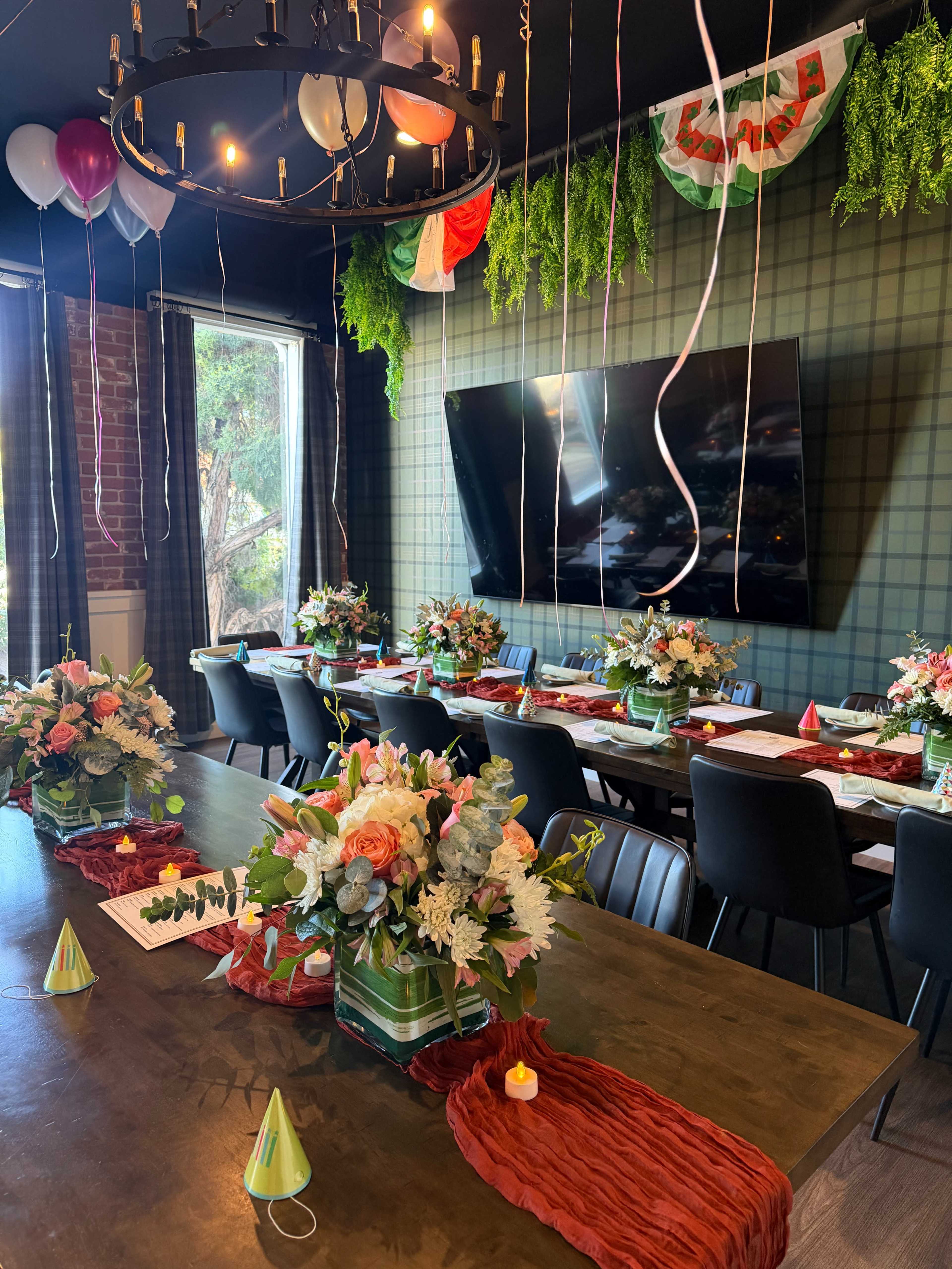 A decorated dining room features a long table set with floral arrangements and festive items, surrounded by chairs and adorned with balloons and streamers.