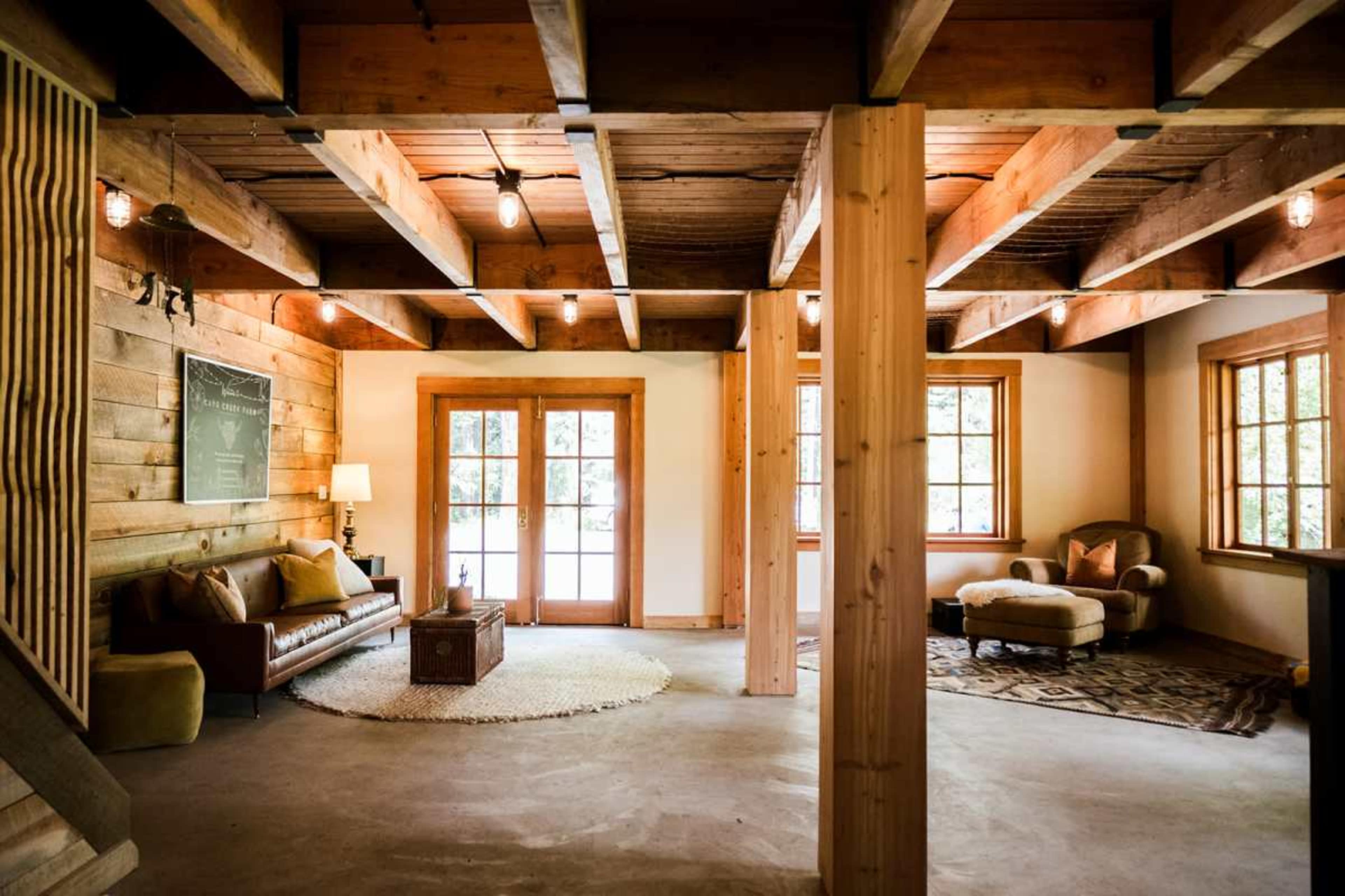 The image depicts a spacious, rustic interior of a cabin with wooden beams, a leather sofa, and large windows allowing natural light to enter.