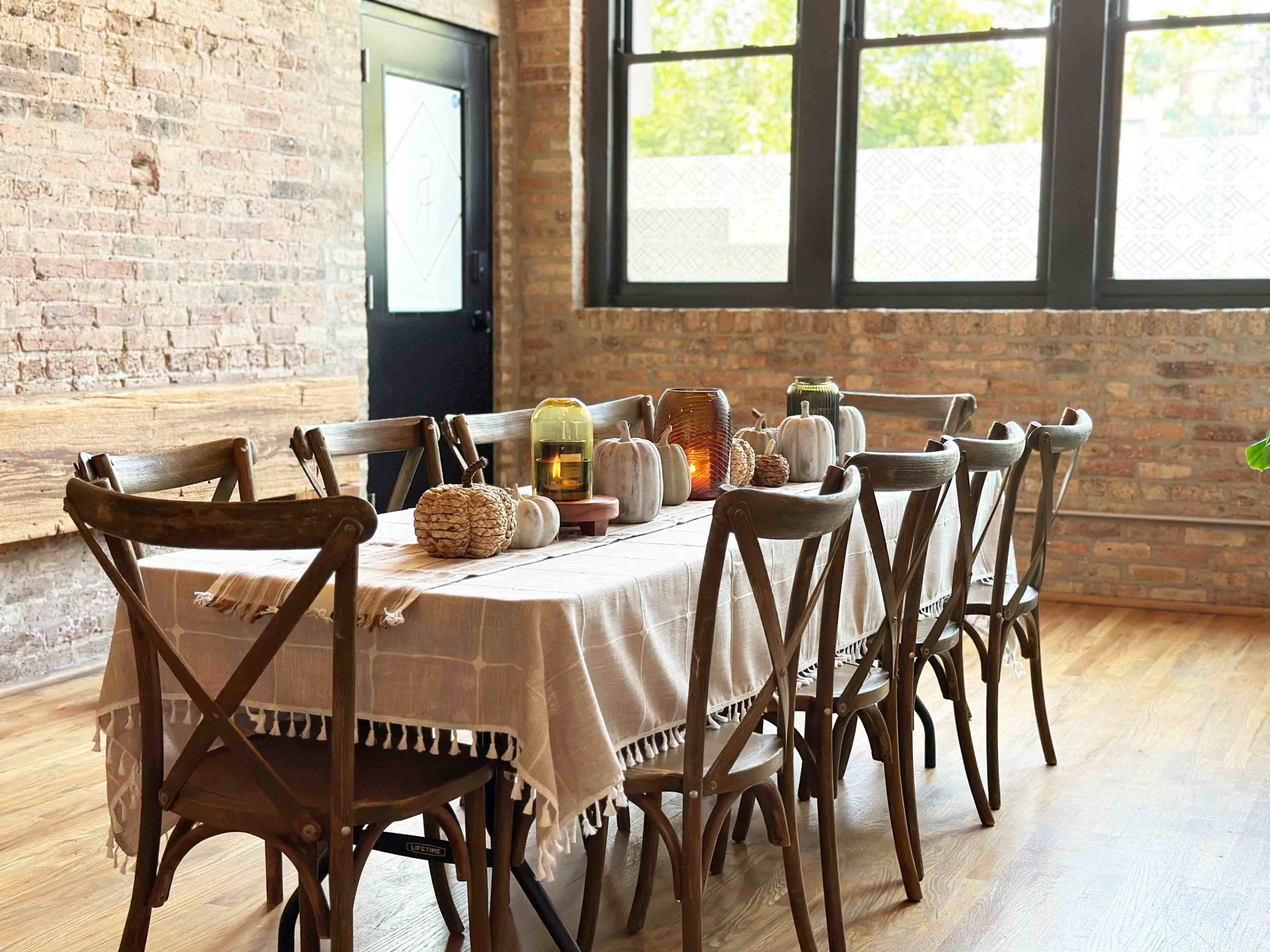 A long wooden dining table with a beige tablecloth is set with various decorative items and surrounded by dark wooden chairs in a sunlit room with brick walls and large windows.