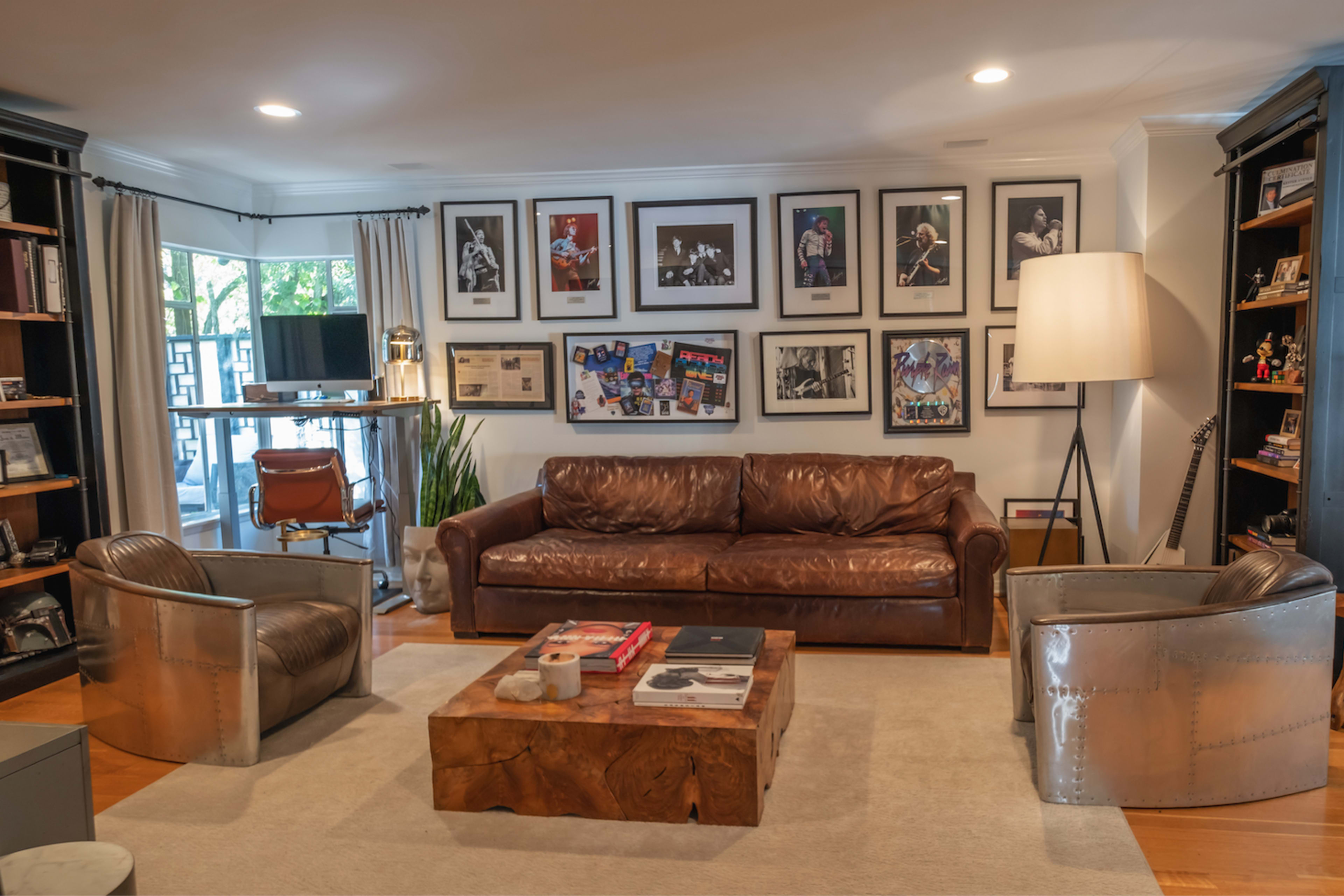 A stylish home office with a brown leather sofa, metal accent chairs, a wooden coffee table, and walls decorated with framed photographs and memorabilia.