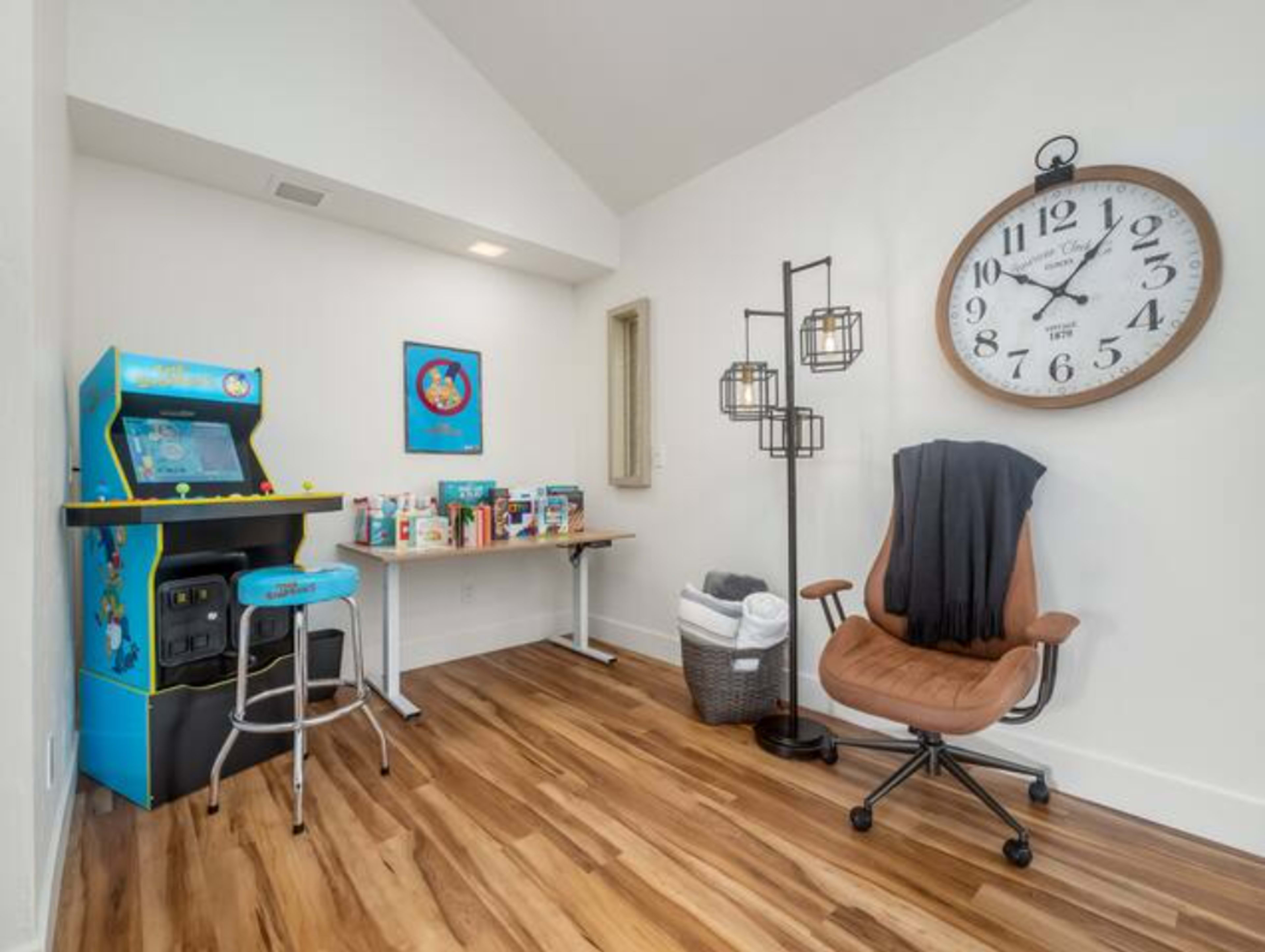 A cozy room featuring a vintage arcade machine, a large wall clock, a brown chair, a table with various boxes, and a laundry basket.