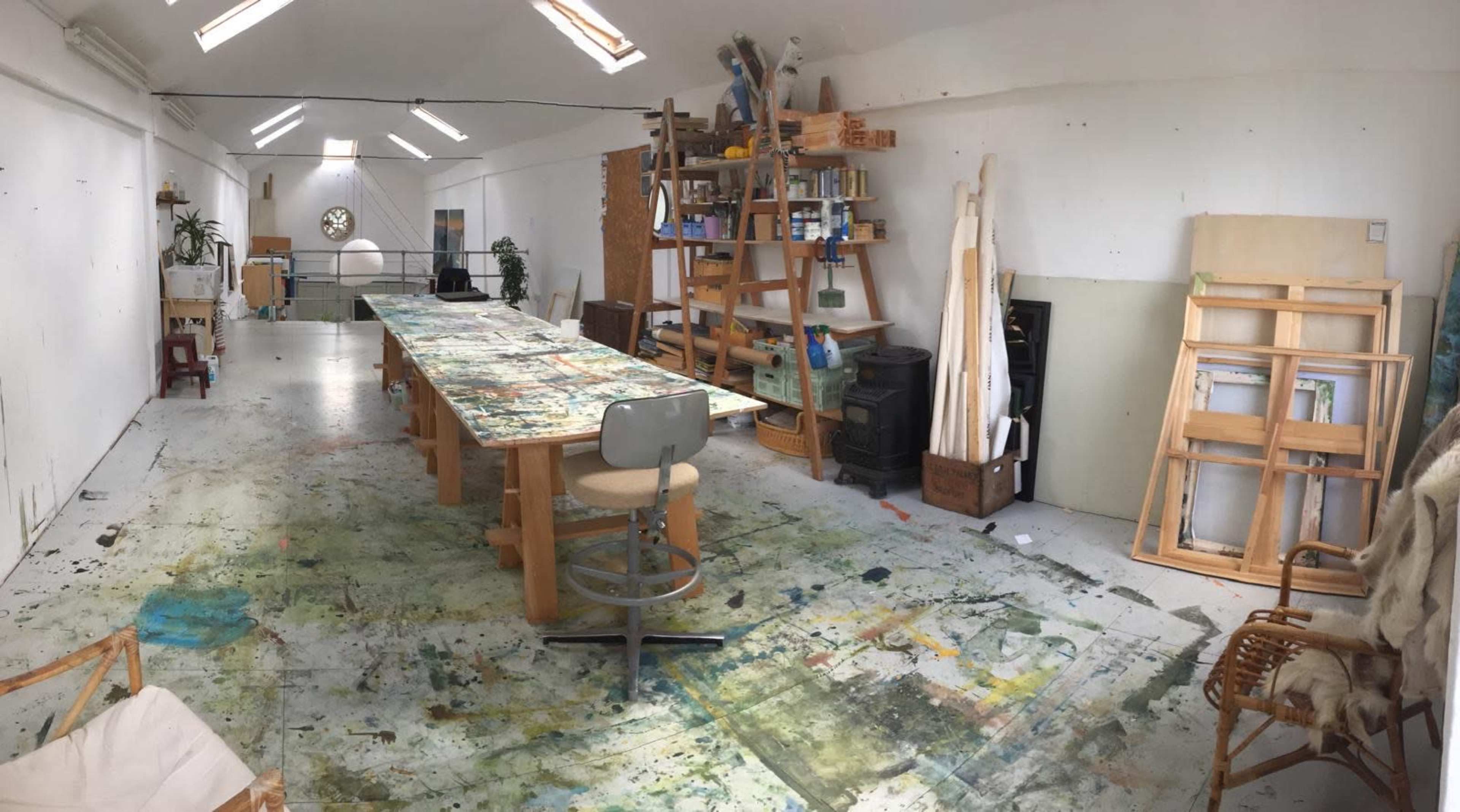 The image shows a spacious art studio with a large table covered in paint splatters, shelves filled with art supplies, and various works in progress along the walls.