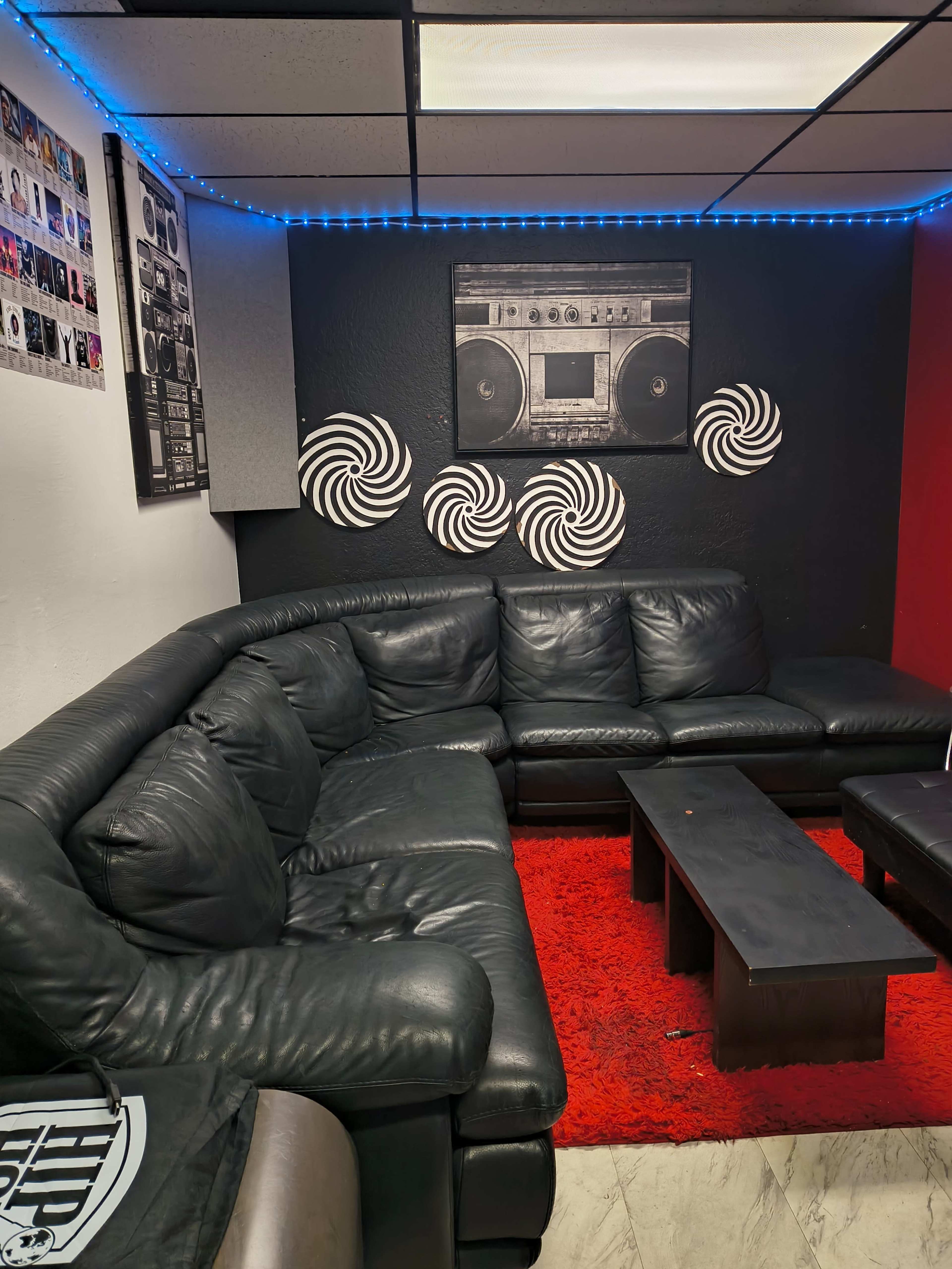 The image shows a lounge area with a black leather sectional sofa, a low coffee table, a red rug, and spiral wall art against a black backdrop.