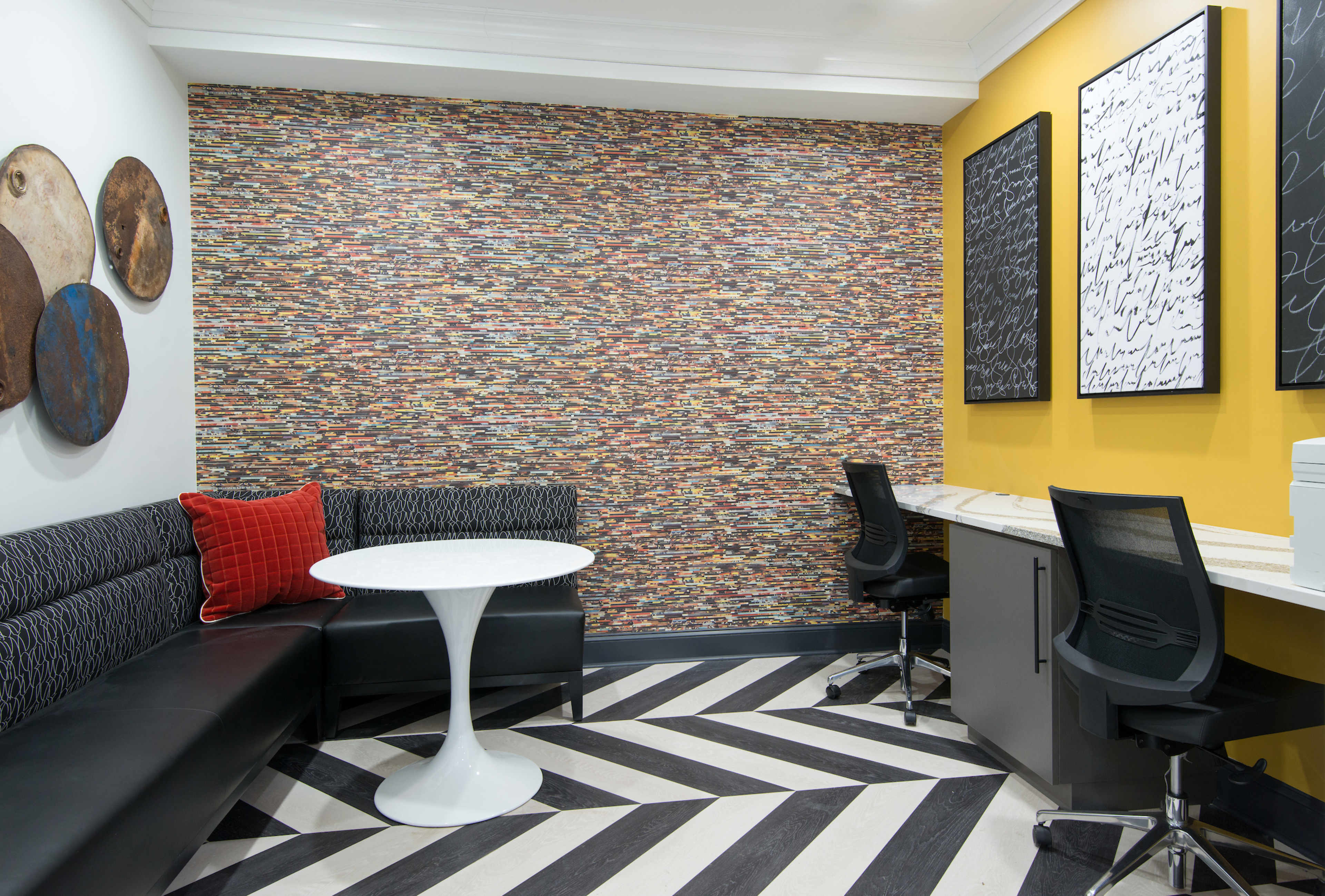 The image shows a modern lounge area with a patterned wall, a round white table, black seating, and two workstations against a yellow wall.