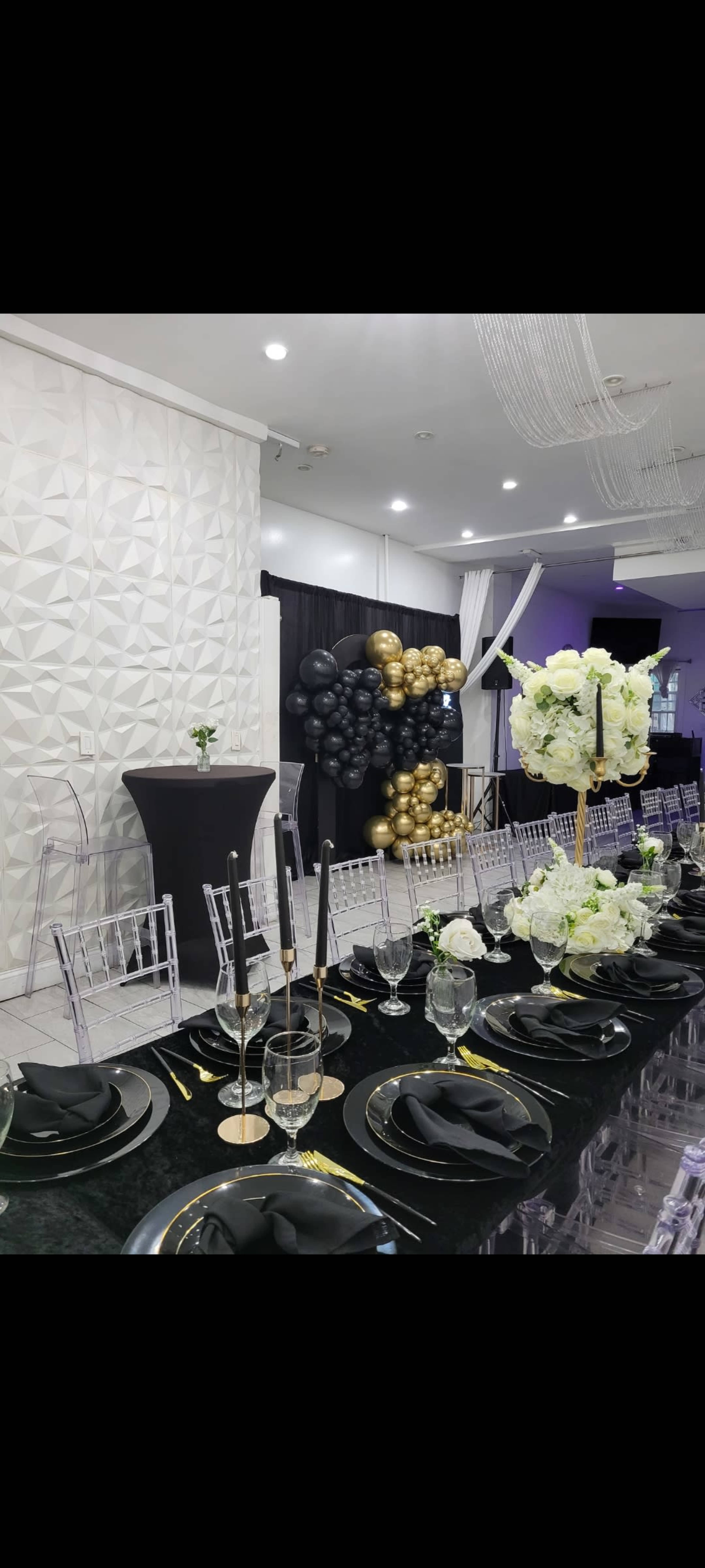 A long, elegantly set dining table features black and gold decor, with clear chairs and floral arrangements, against a backdrop of a balloon arch and geometric wall.