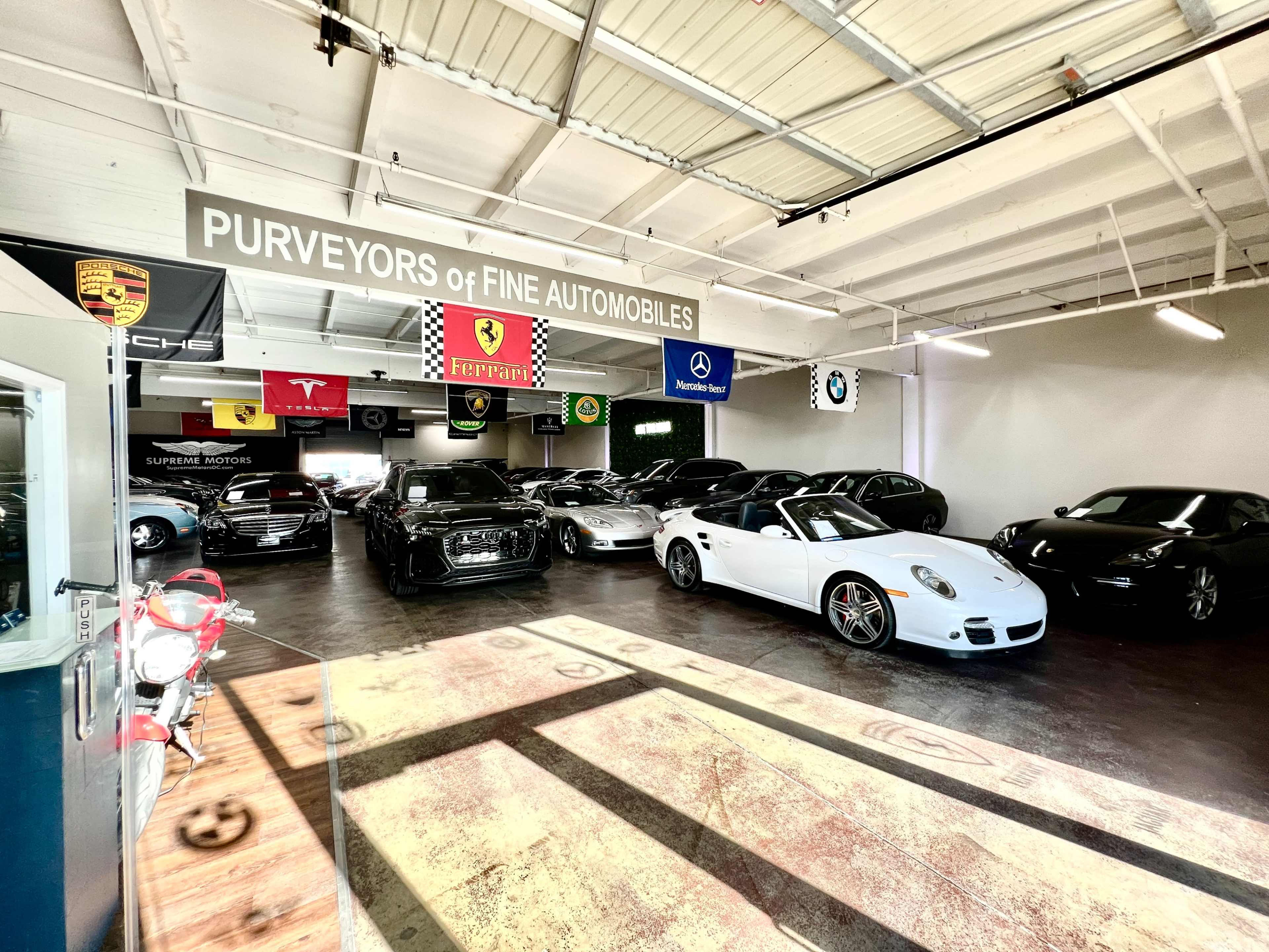 The image shows an indoor luxury car showroom featuring several high-end vehicles, including sports cars and sedans, with branded banners hanging from the ceiling.