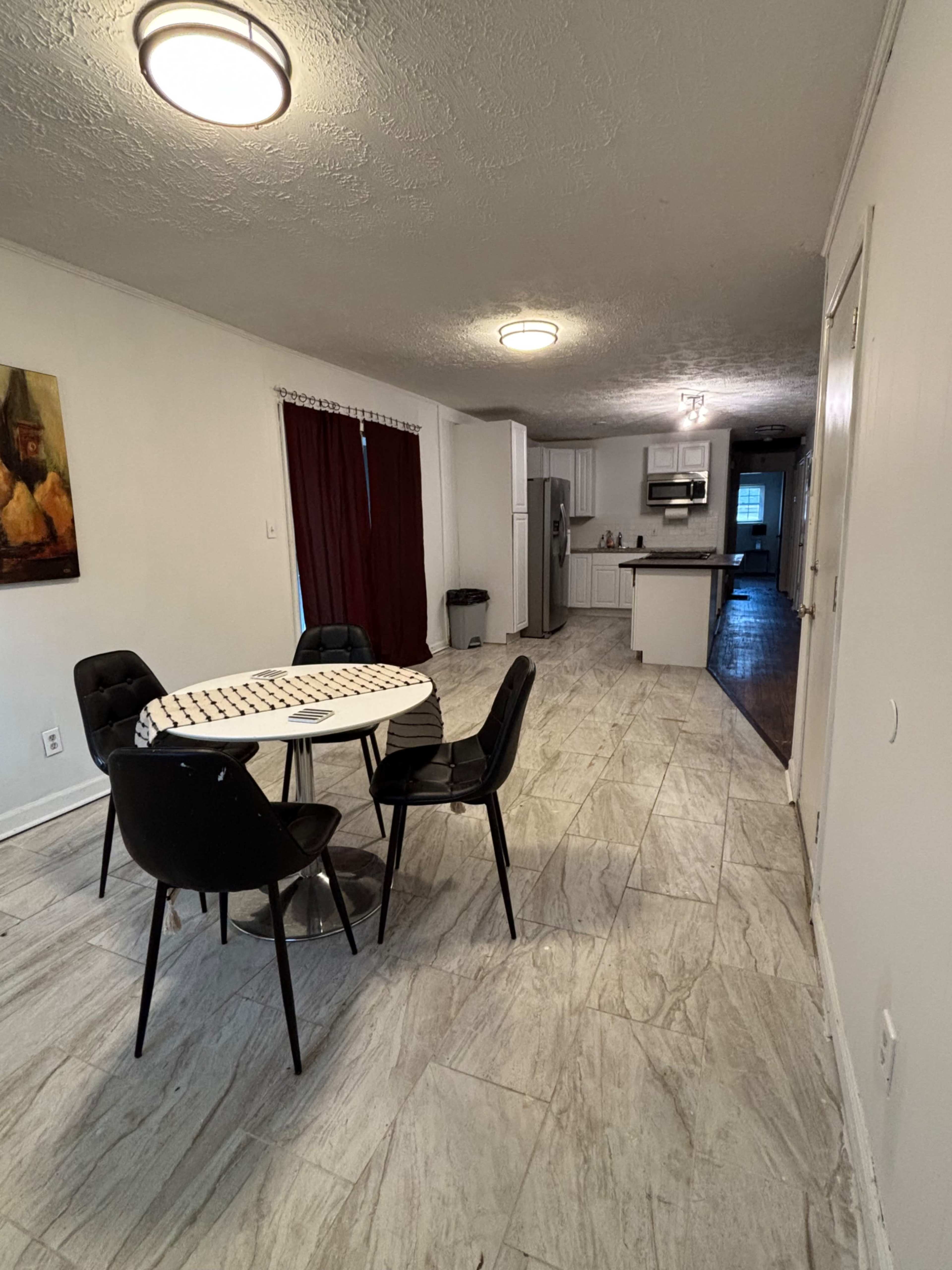 The image shows a dining area with a round table and four black chairs, leading to a kitchen in the background.