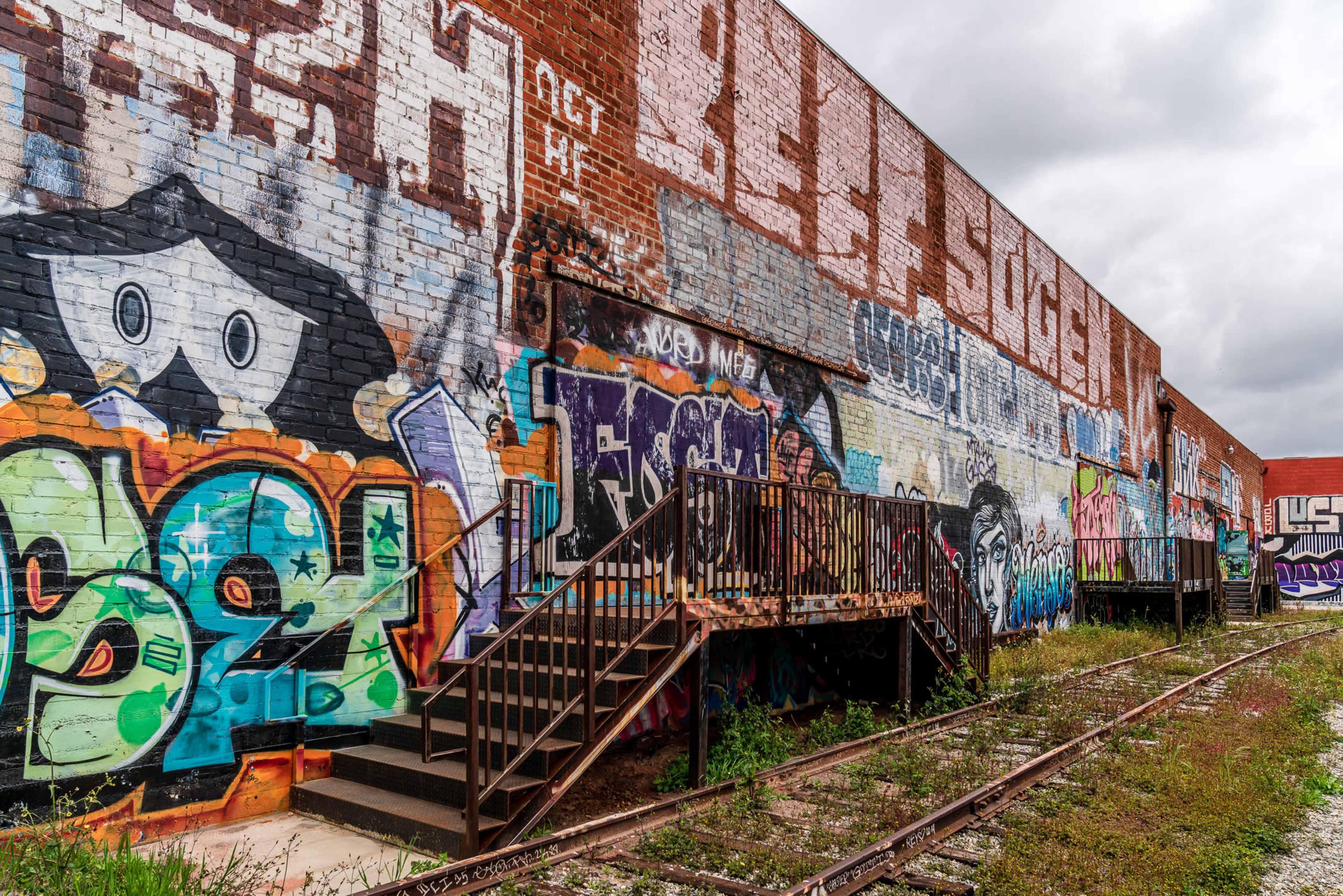 A brick wall covered with colorful graffiti and murals stands beside a set of metal stairs leading to a platform, with overgrown grass and an abandoned railway track in the foreground.