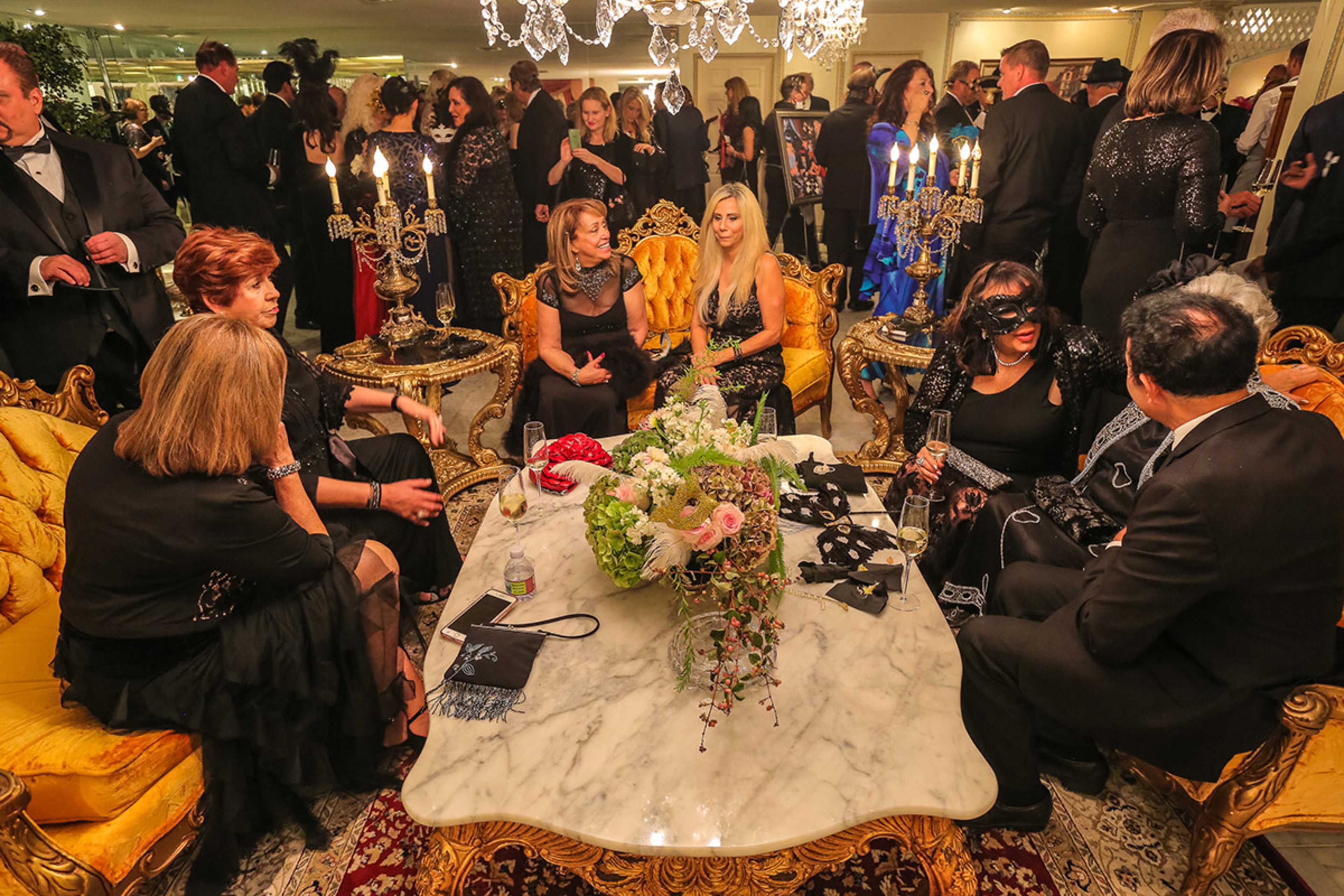 A group of elegantly dressed individuals socializes around a marble table in a lavishly decorated room.