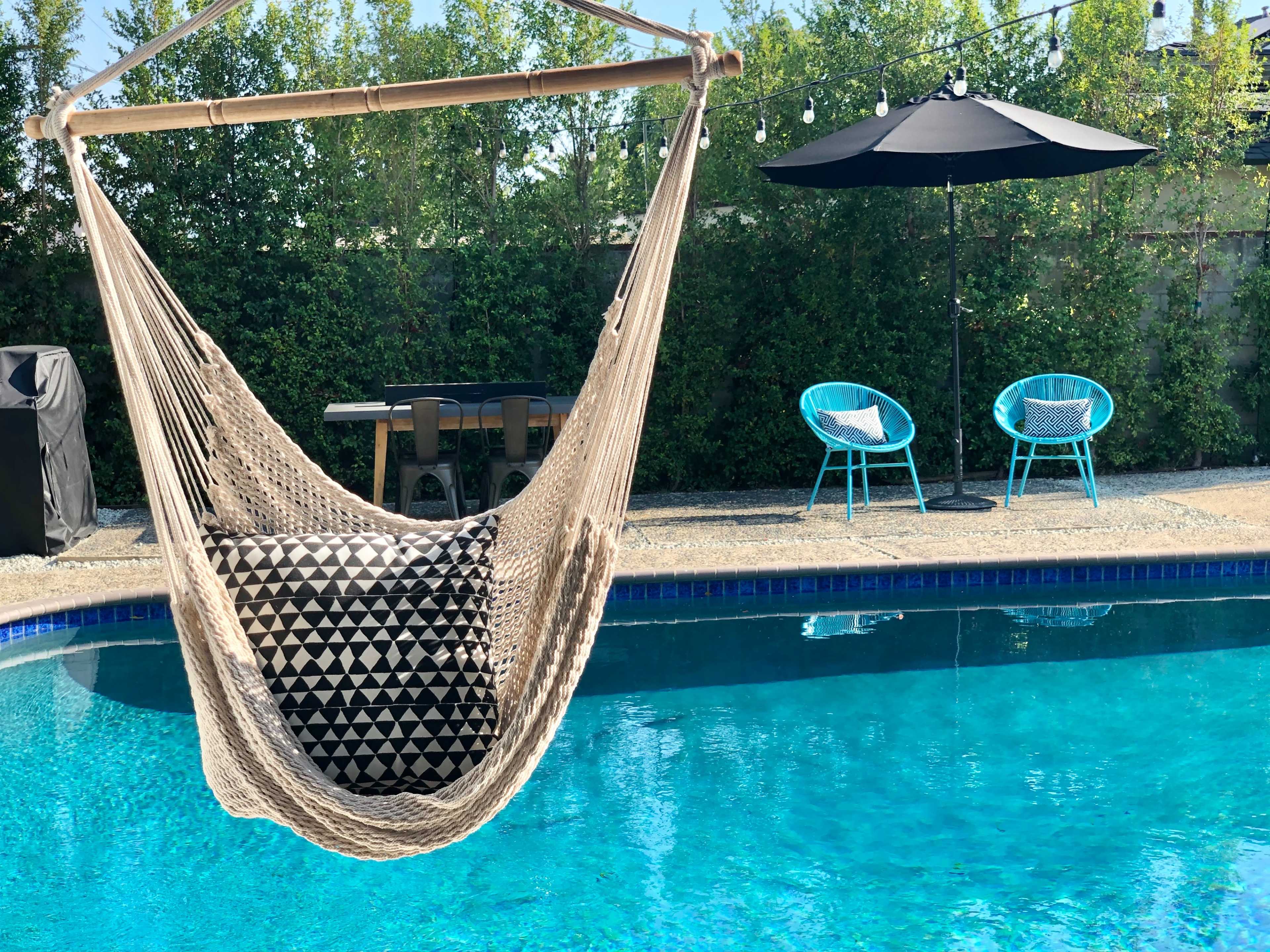 A hammock is suspended near a swimming pool surrounded by green foliage, with two blue chairs and a table nearby.