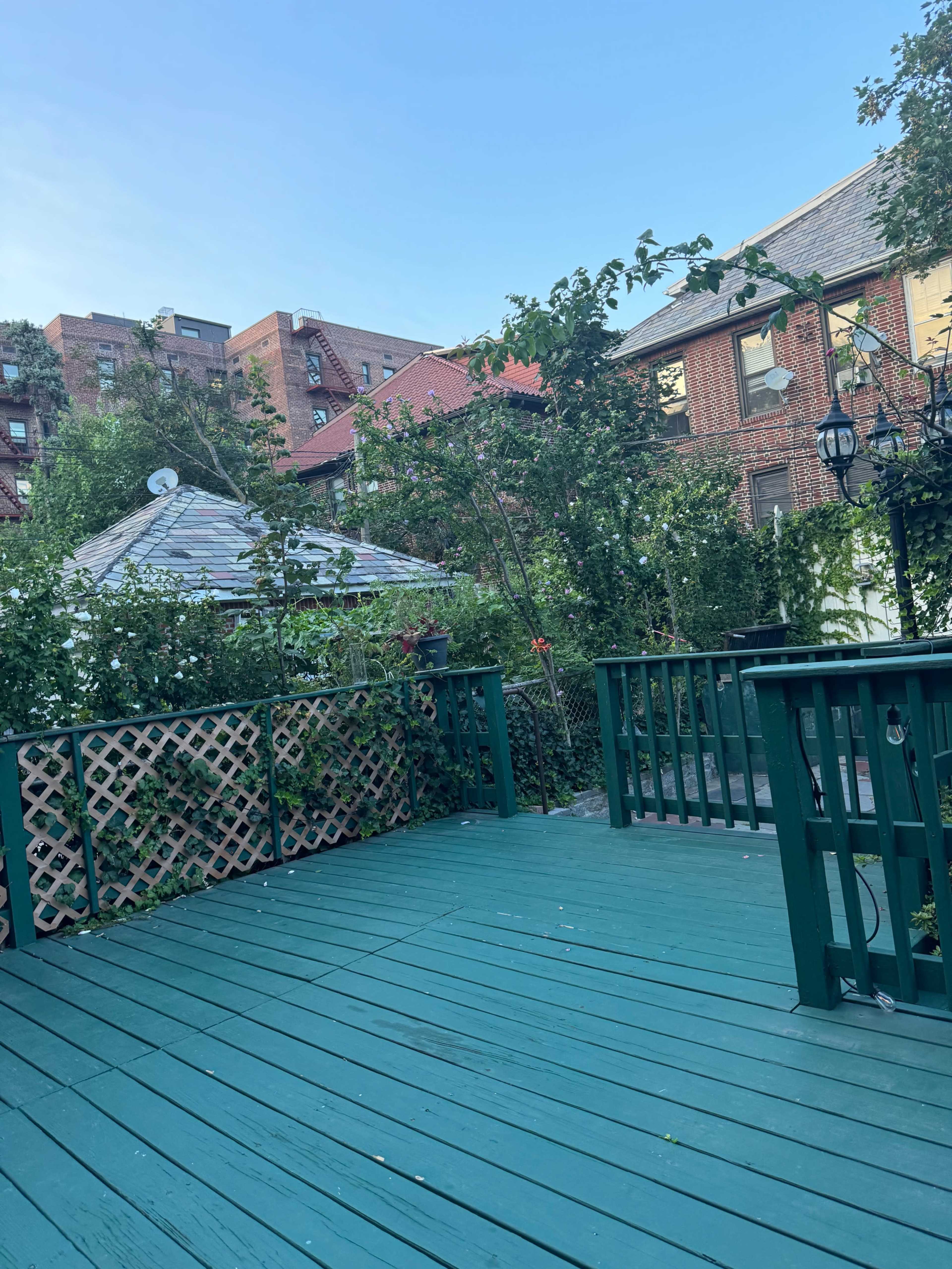 The image shows a green wooden deck surrounded by lush greenery and urban buildings in the background.