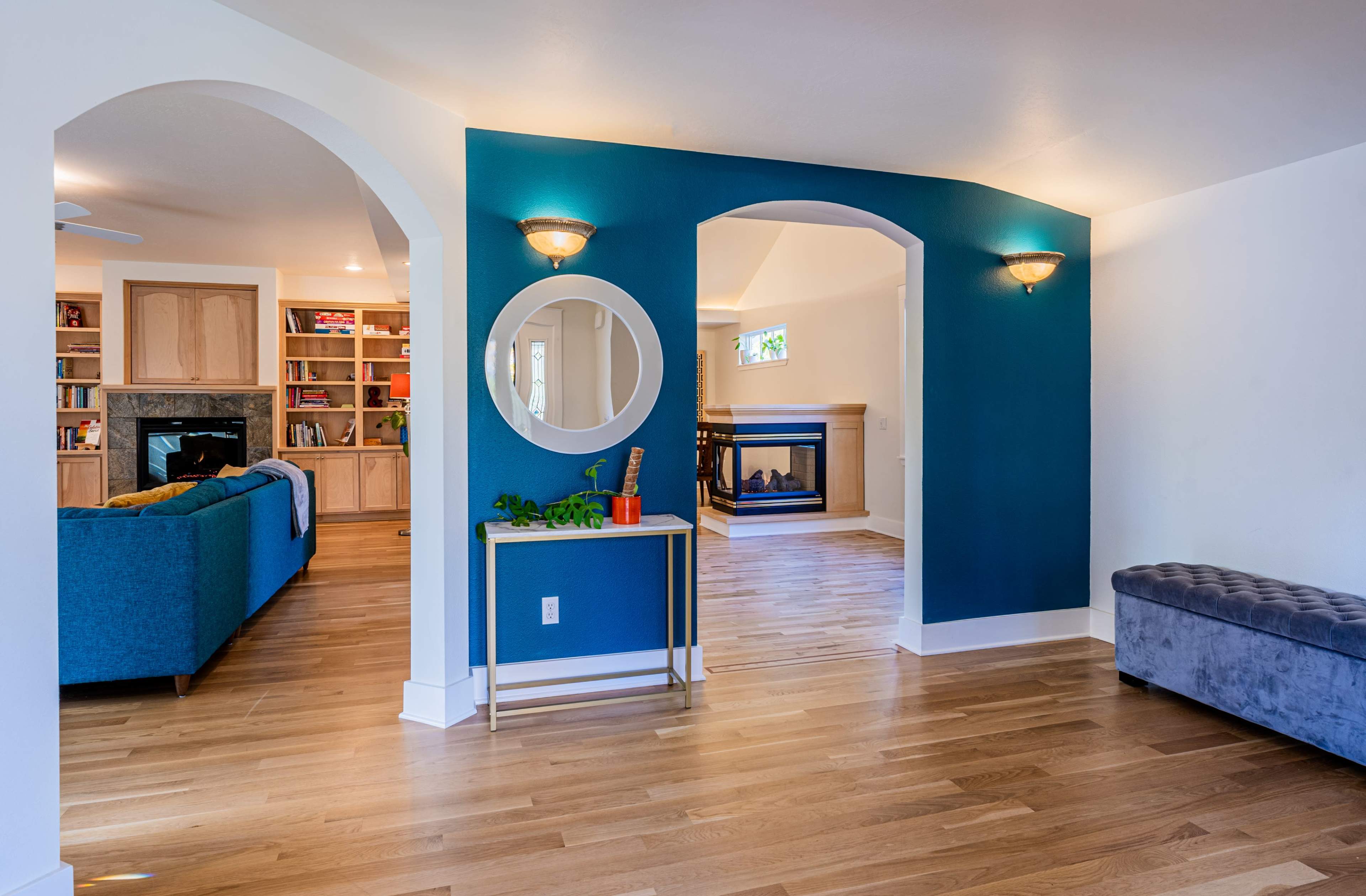 The image shows a modern interior with a blue accent wall, a round mirror, and a cozy seating area visible beyond an archway.