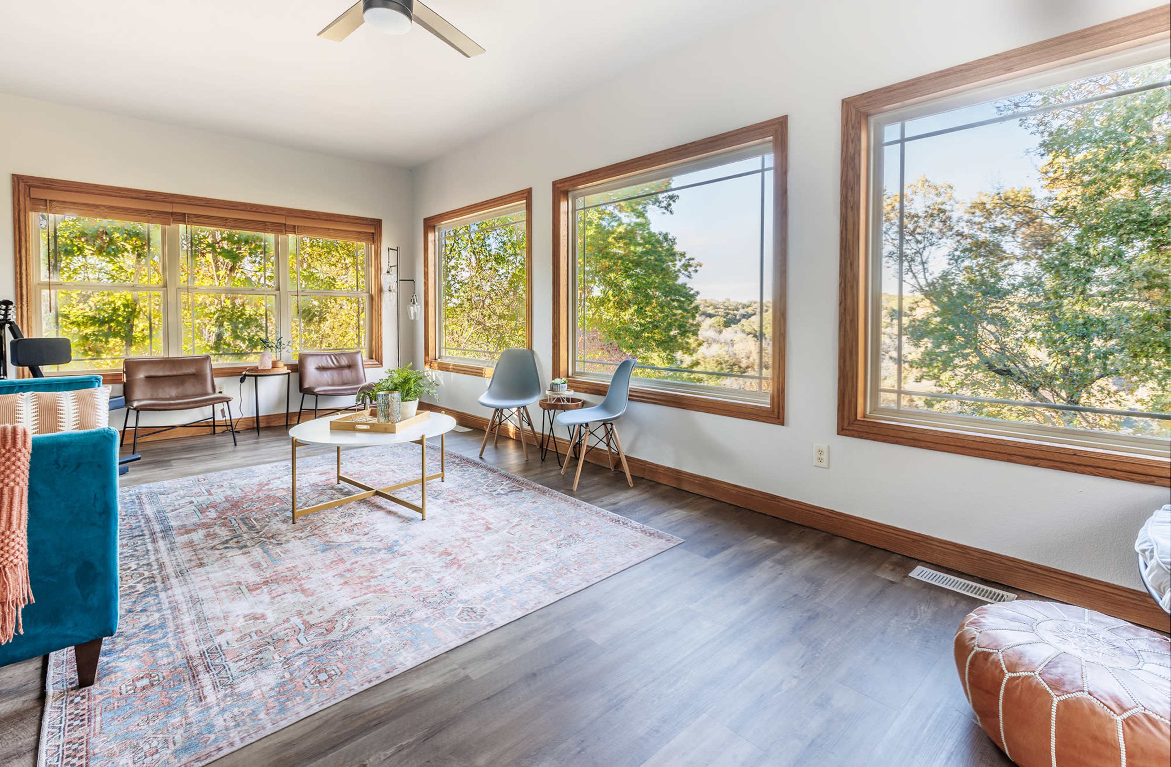 The room features large windows that provide a view of trees, with a seating area consisting of a sofa, chairs, and a coffee table on a patterned rug.