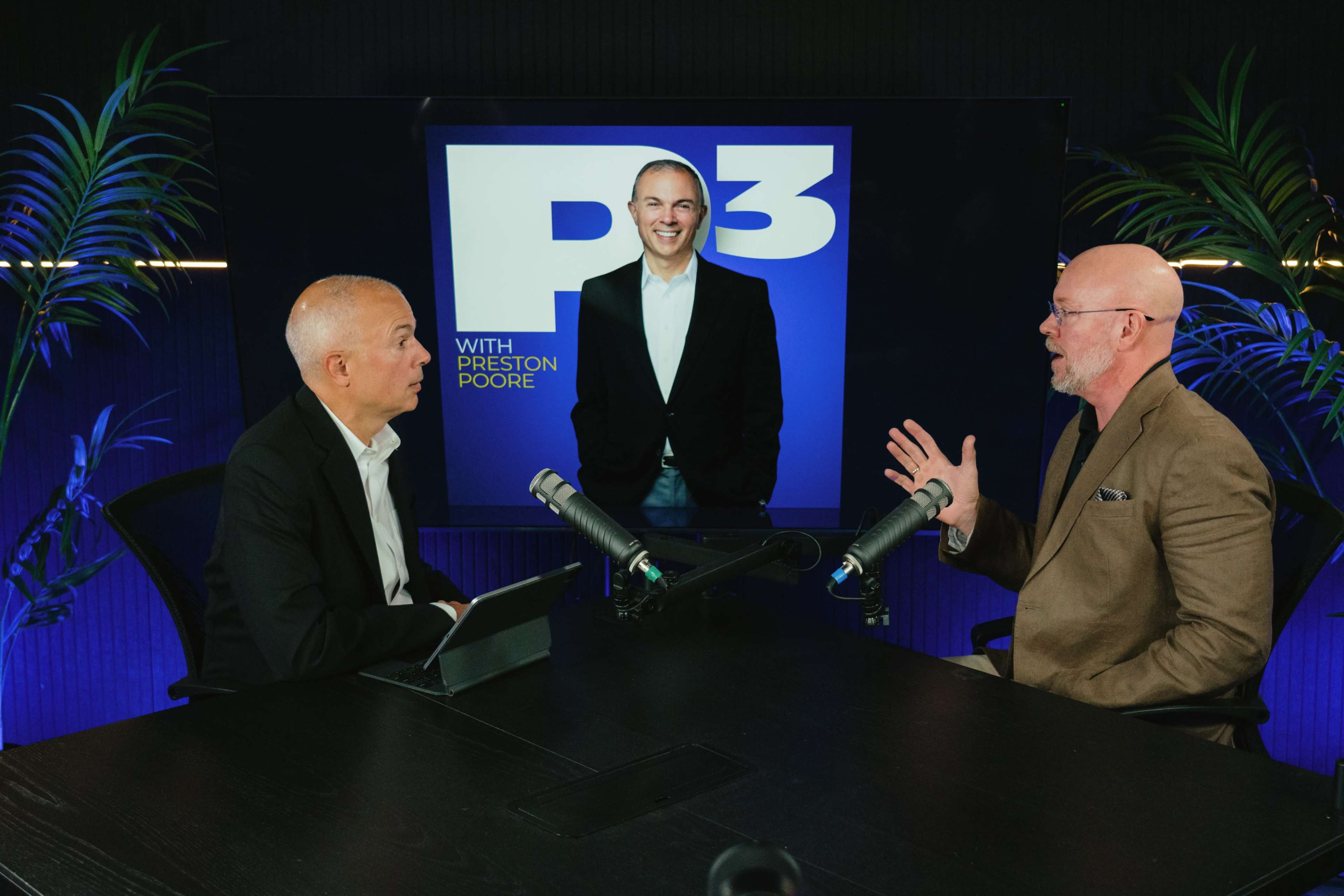 Two men are seated at a table with microphones, engaged in conversation, while a third man stands in front of a large screen displaying the logo "P3 with Preston Poore."