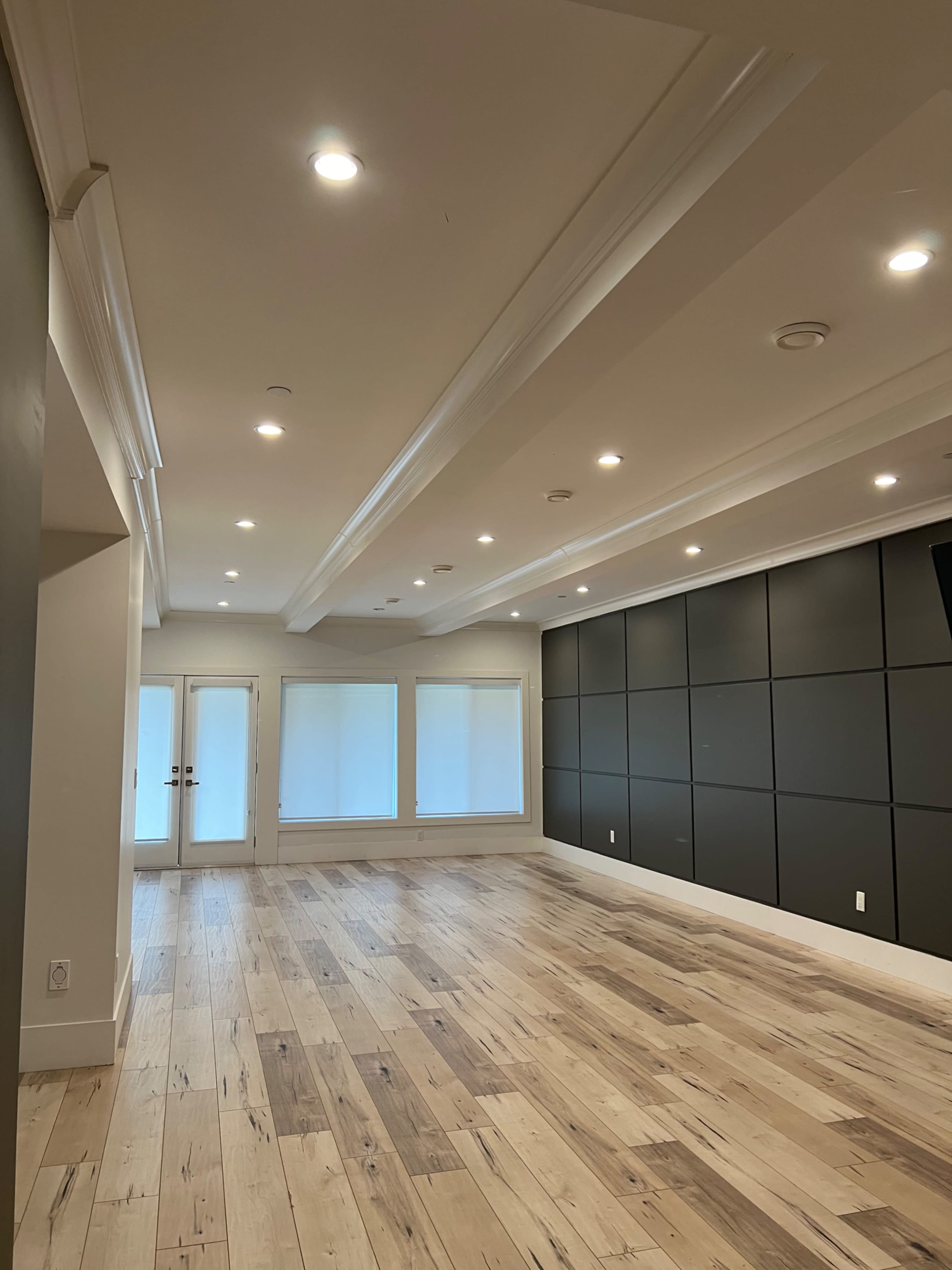 A spacious room with wooden flooring, recessed lighting, and large windows covered by shades.