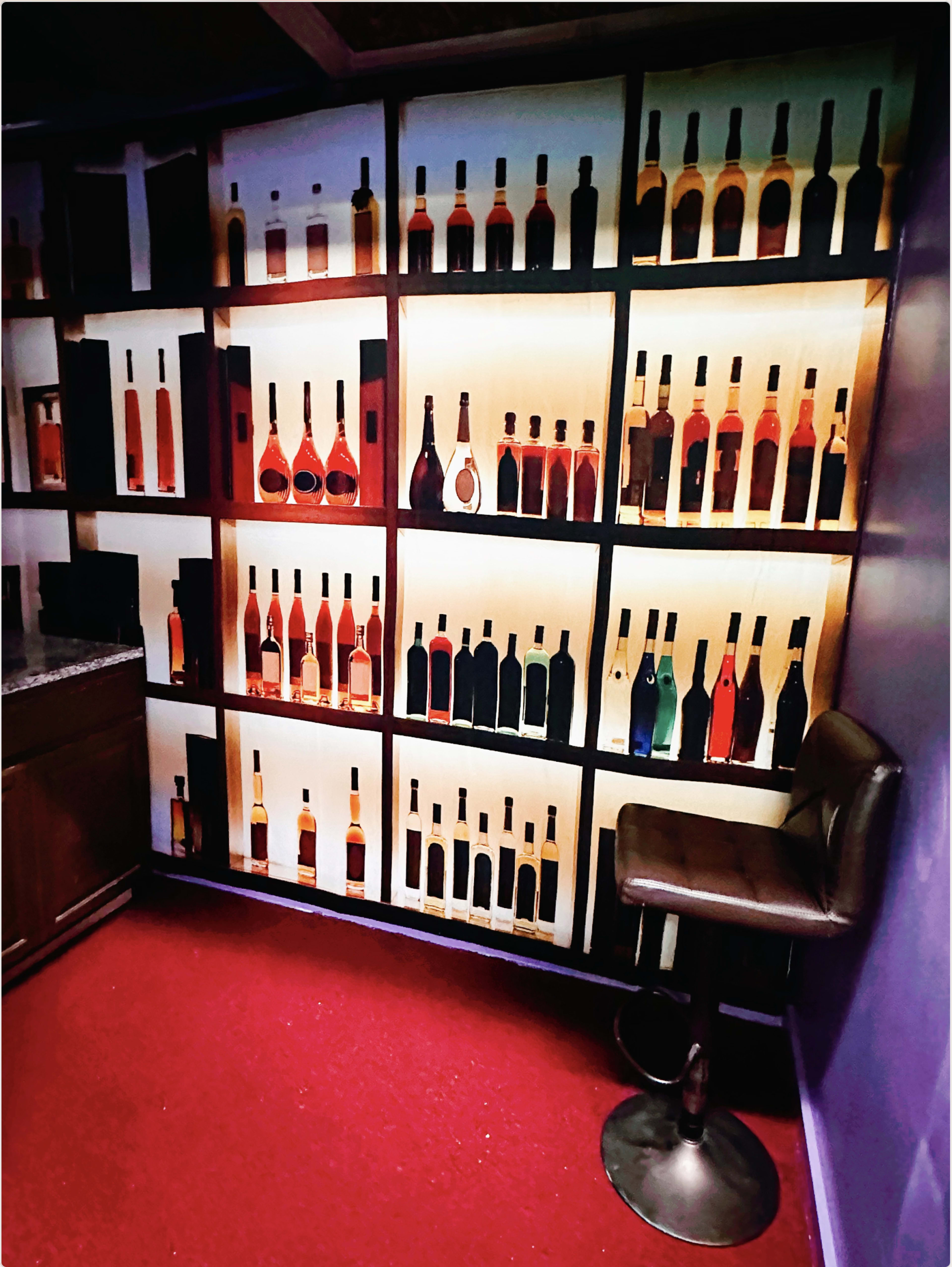 A wall with a backlit display of various bottle silhouettes against a purple backdrop, accompanied by a bar stool on a red carpeted floor.