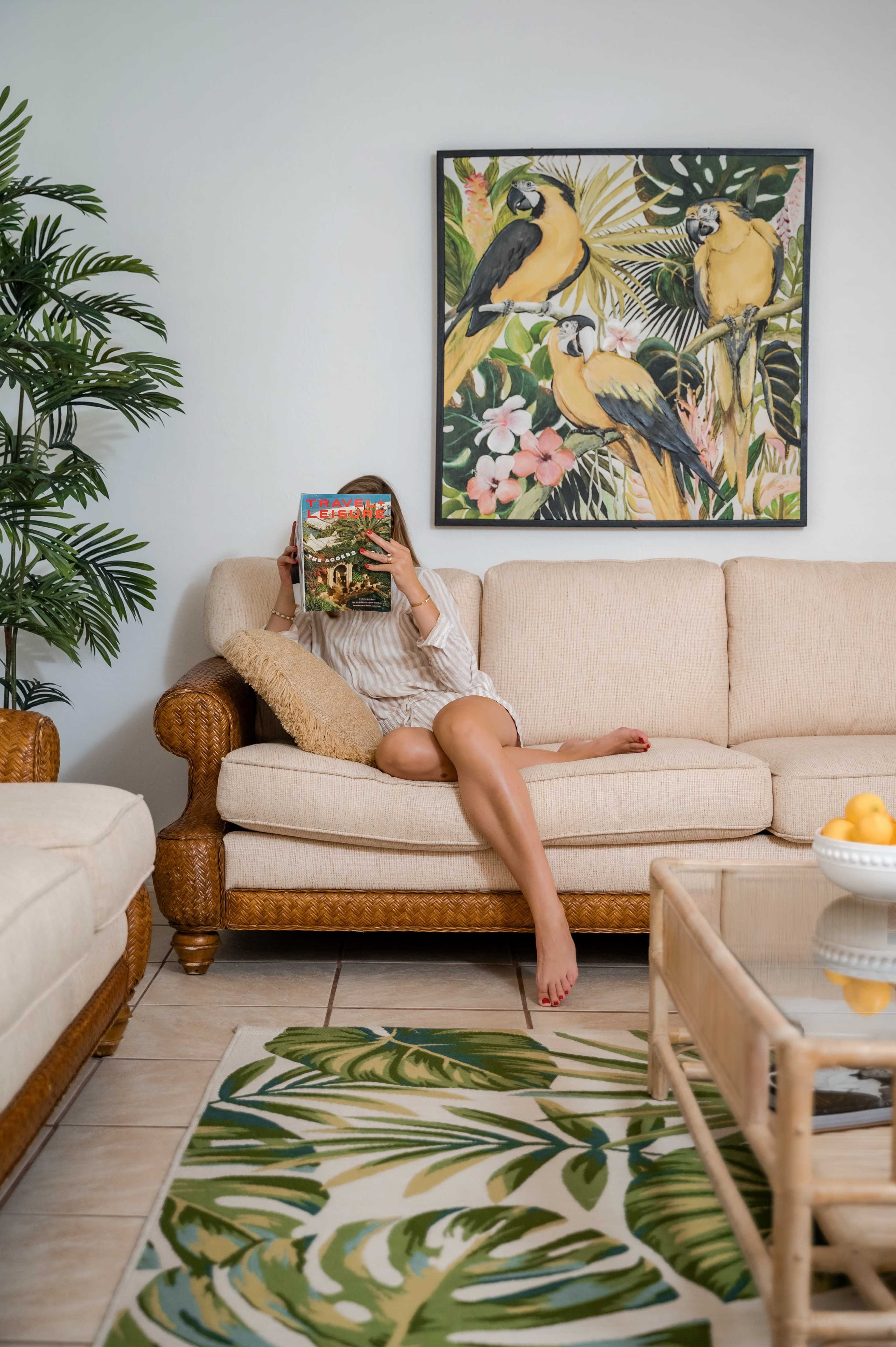 A woman sits on a beige sofa, holding a magazine in front of her face, while a colorful bird-themed artwork hangs on the wall behind her.
