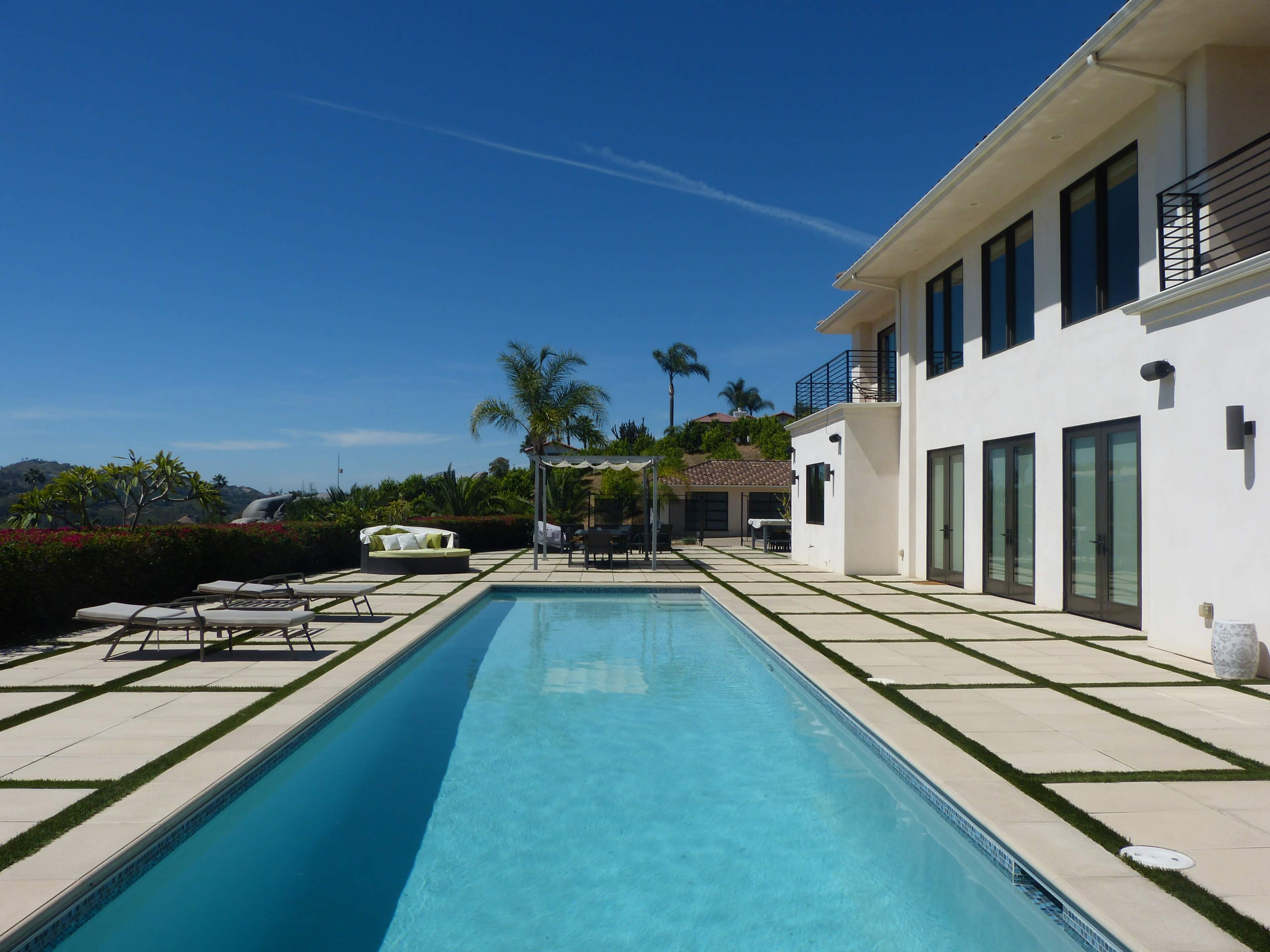 The image shows a modern home with a swimming pool surrounded by lounge chairs and a pergola, set against a clear blue sky.