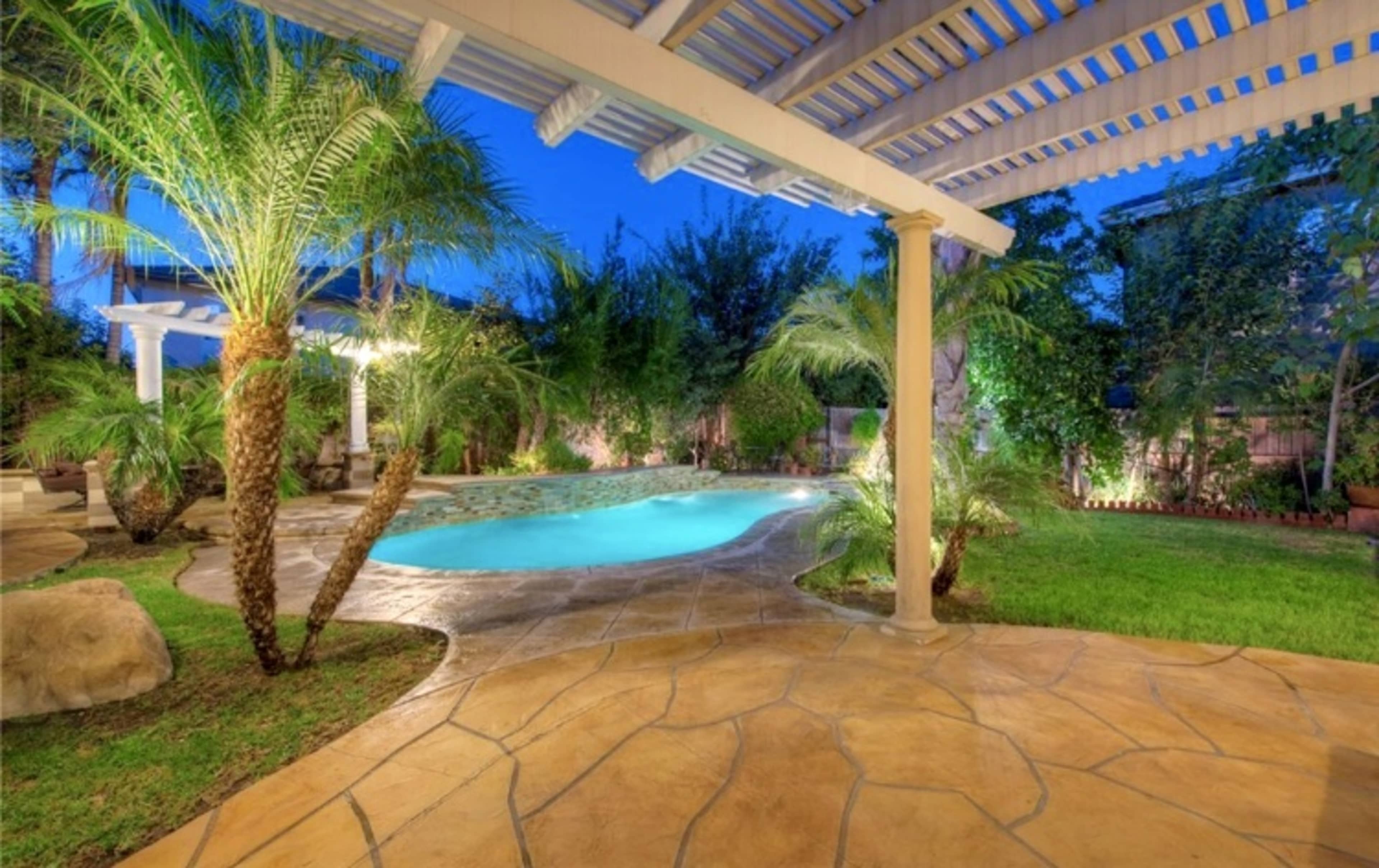 The image shows a landscaped backyard with a swimming pool surrounded by palm trees and a stone patio under a pergola.