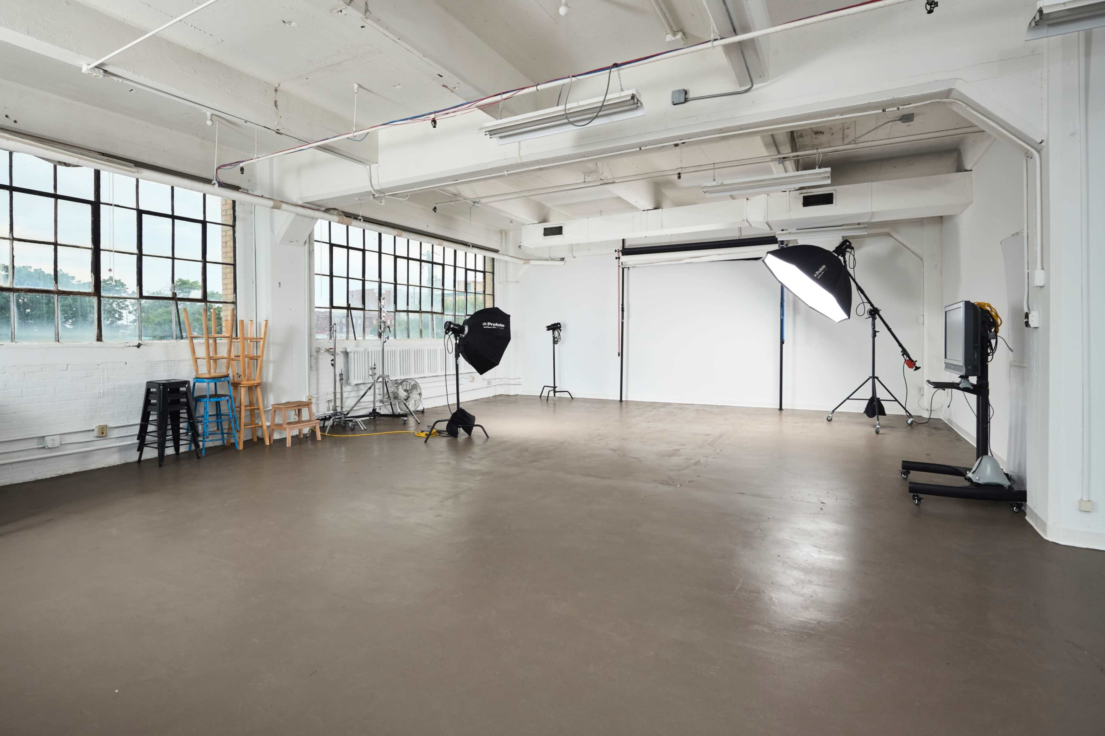 The image shows a spacious photography studio with large windows, a seamless backdrop, and various lighting equipment arranged throughout the room.