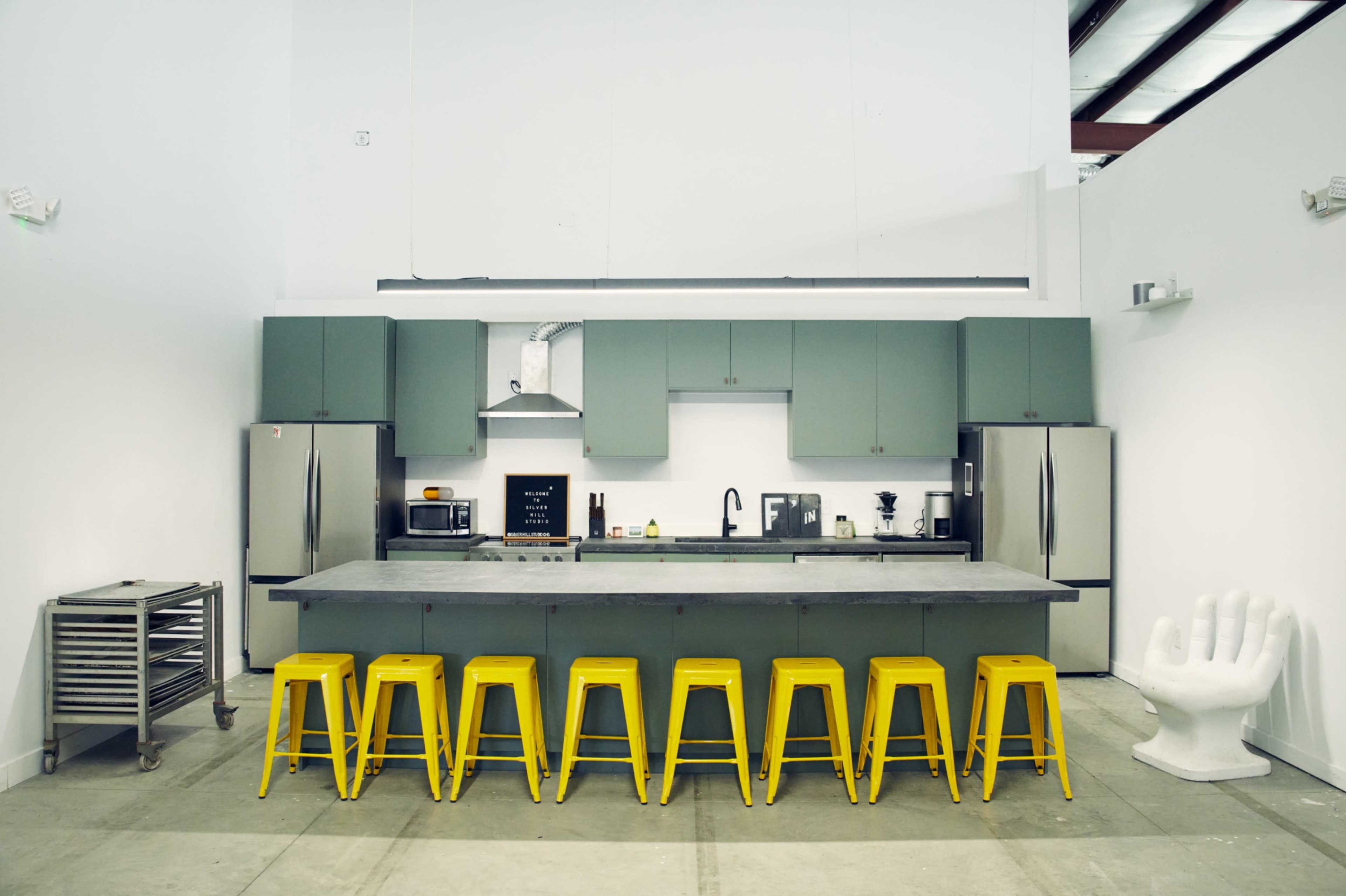 The image shows a modern kitchen with gray cabinets, stainless steel appliances, and a long concrete island with yellow stools arranged in front of it.