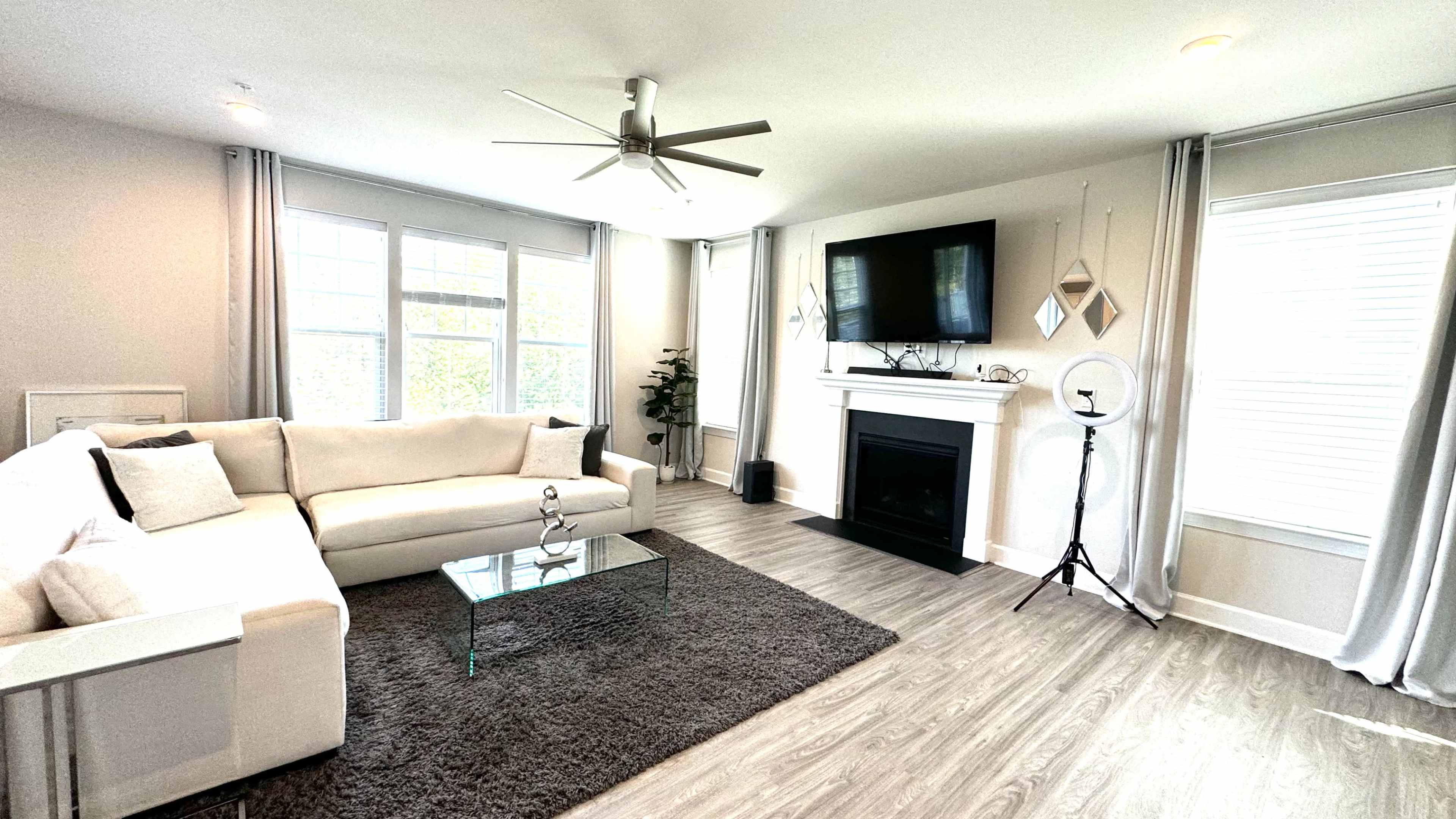 The living room features a white sectional sofa, a glass coffee table, a fireplace with a television above, and large windows with light curtains.