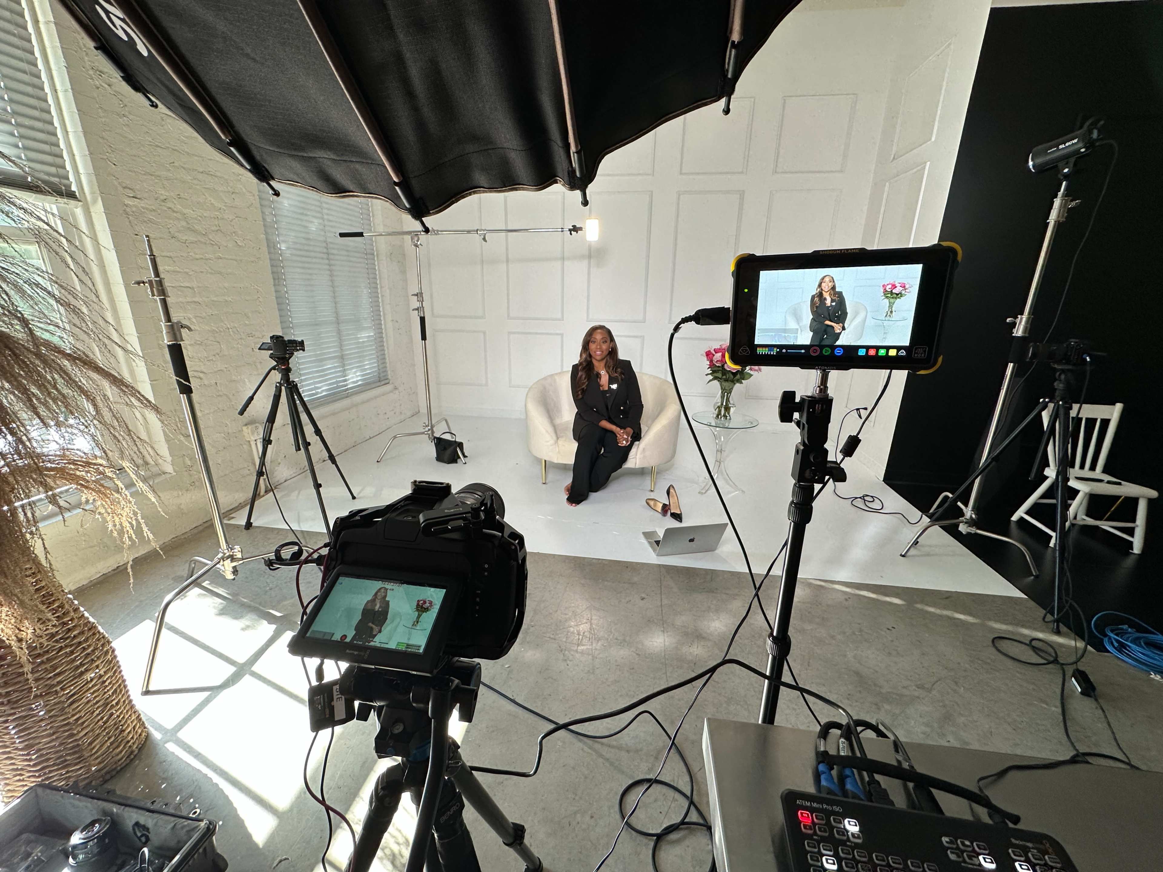 A woman sits on a white chair in a well-lit studio surrounded by multiple cameras and lighting equipment.