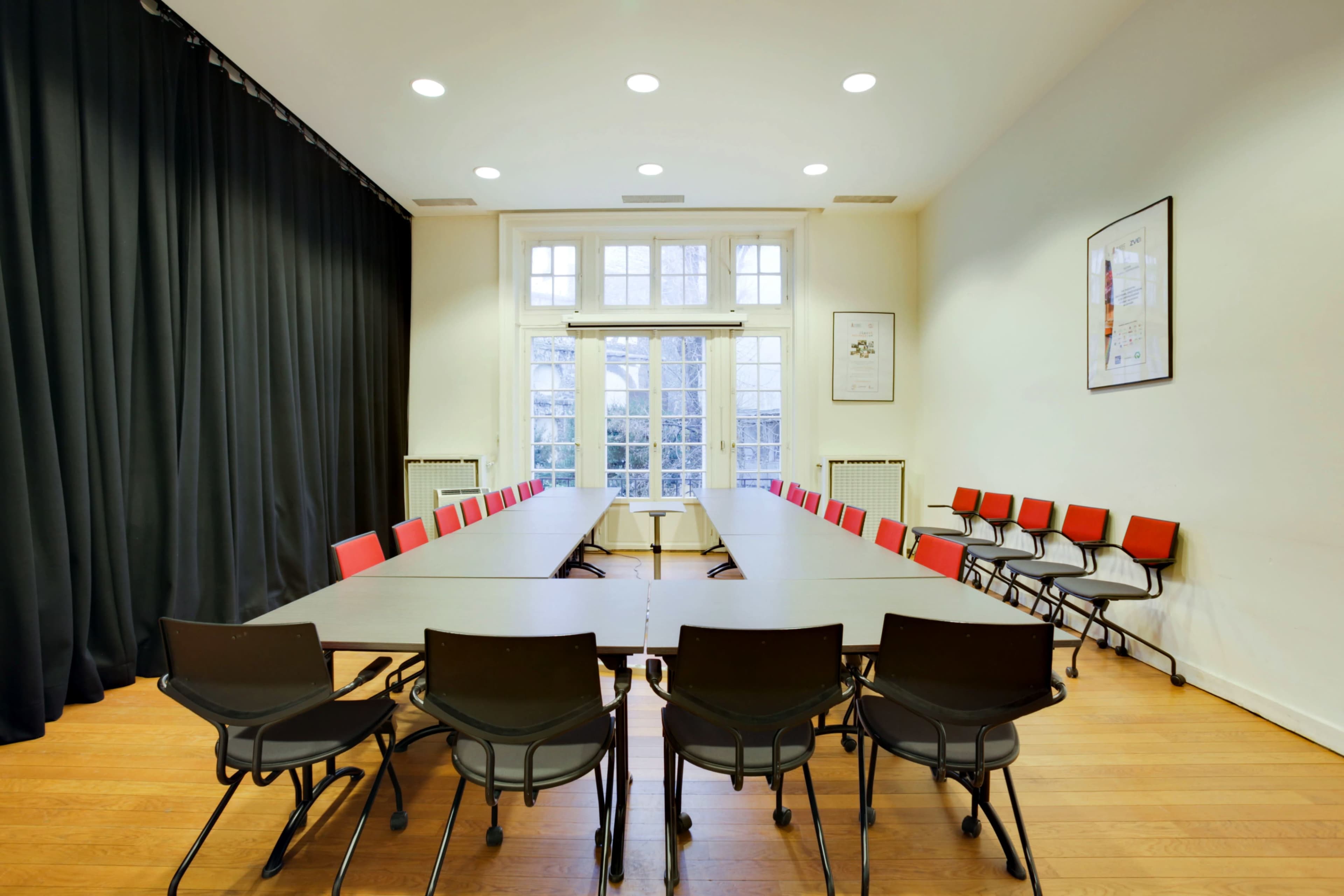 A spacious meeting room features a long table surrounded by black and red chairs, with large windows providing natural light.