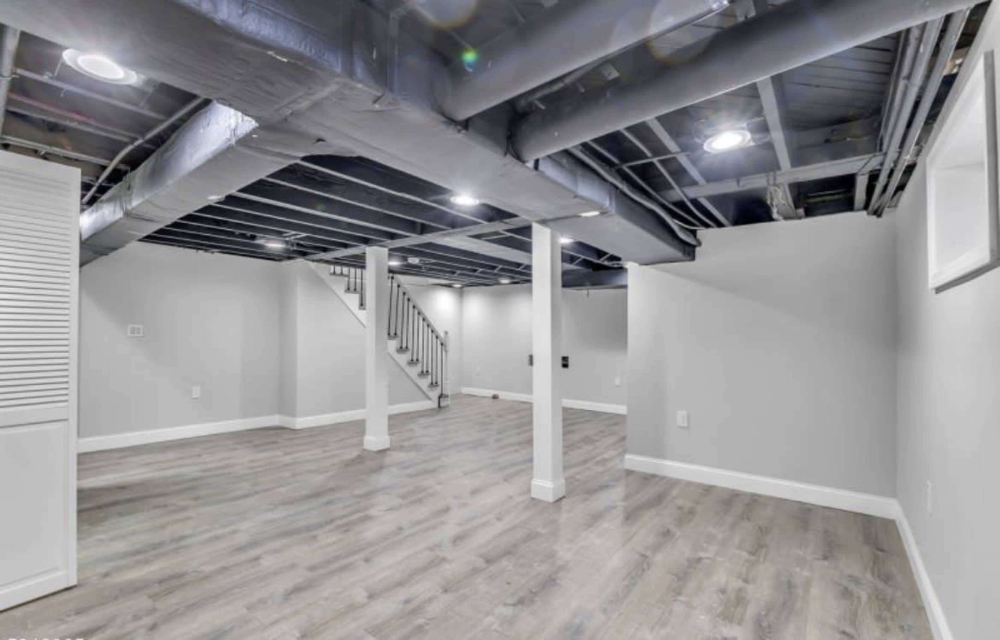 The image shows a partially finished basement with light-colored walls, wooden flooring, and exposed ceiling beams.