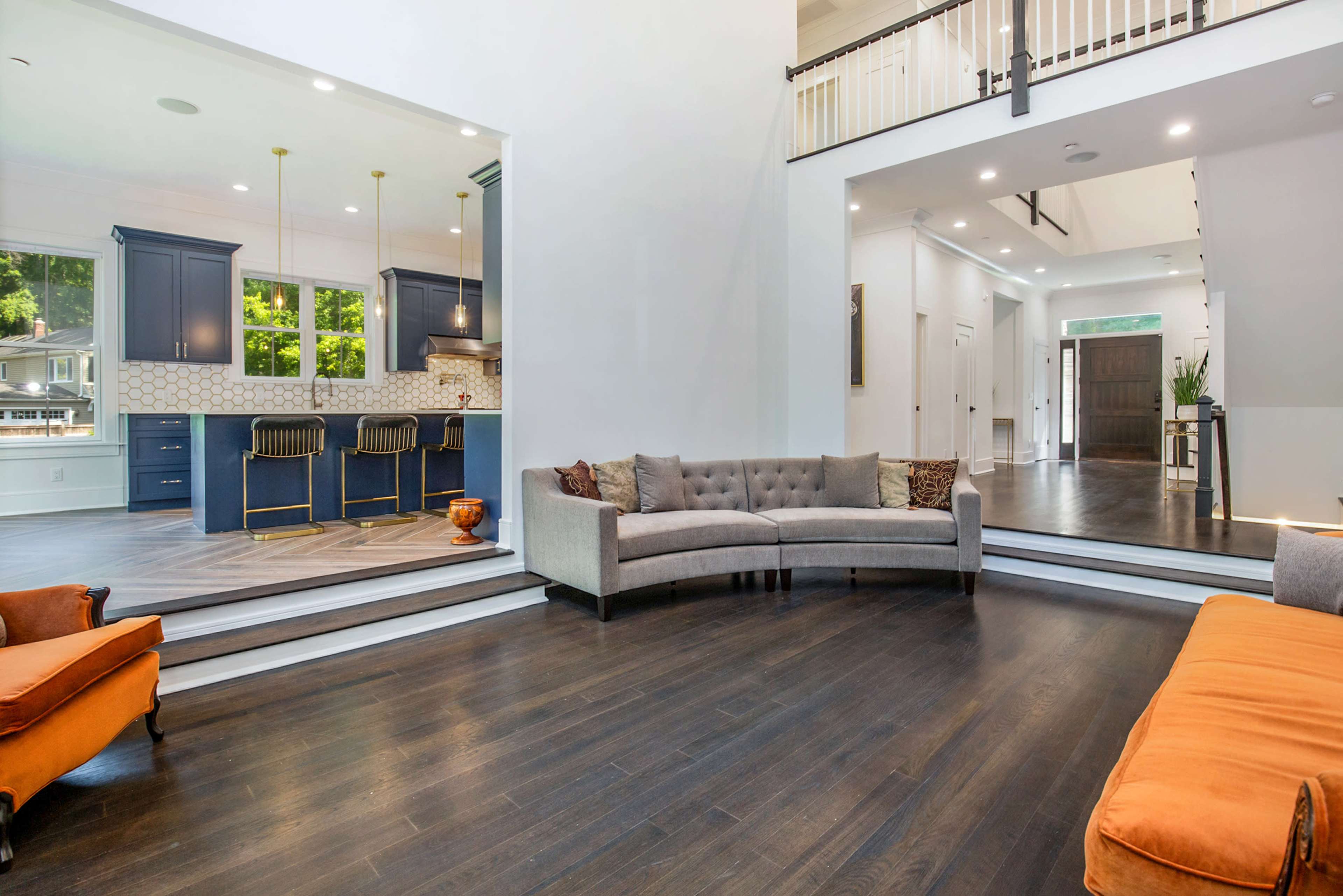 A modern living space featuring a gray sectional sofa, a bright orange couch, and a stylish kitchen with dark blue cabinetry and pendant lighting.