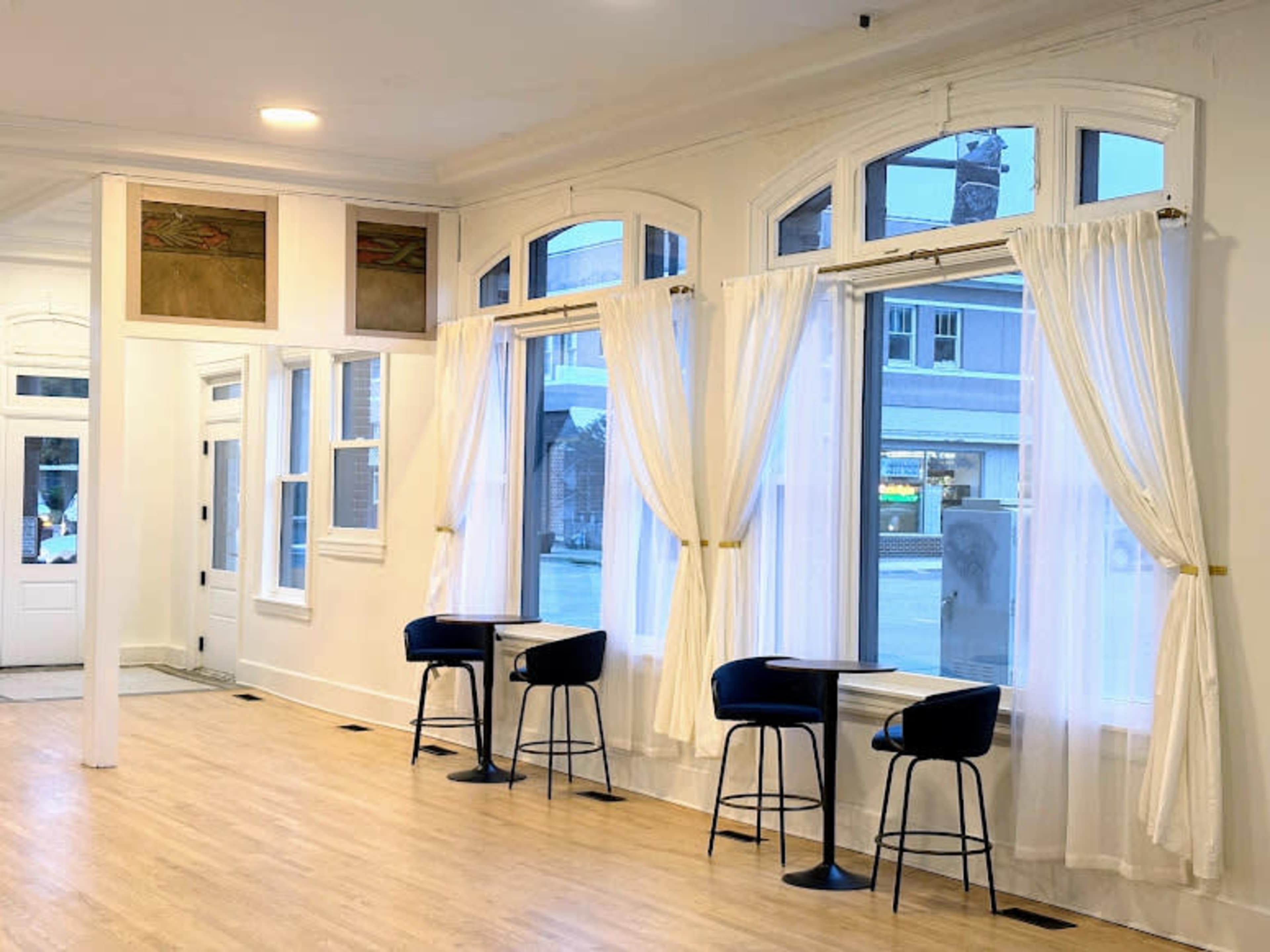 The image shows a modern, well-lit interior space featuring tall windows, light wooden flooring, and two high stools positioned near the windows.