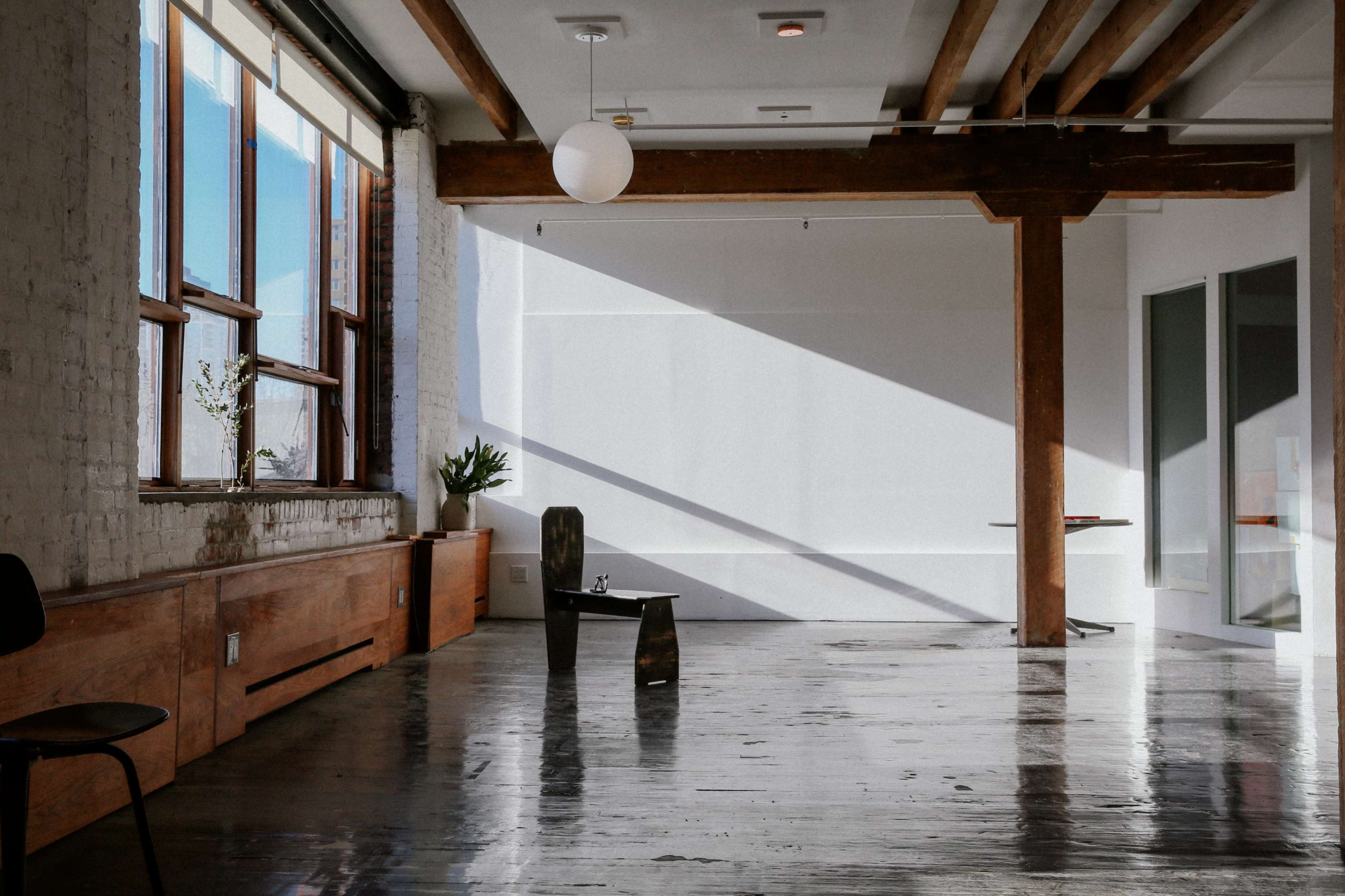 The image shows a bright, open room with high ceilings, wooden beams, large windows, and polished floors, featuring minimalist furniture.