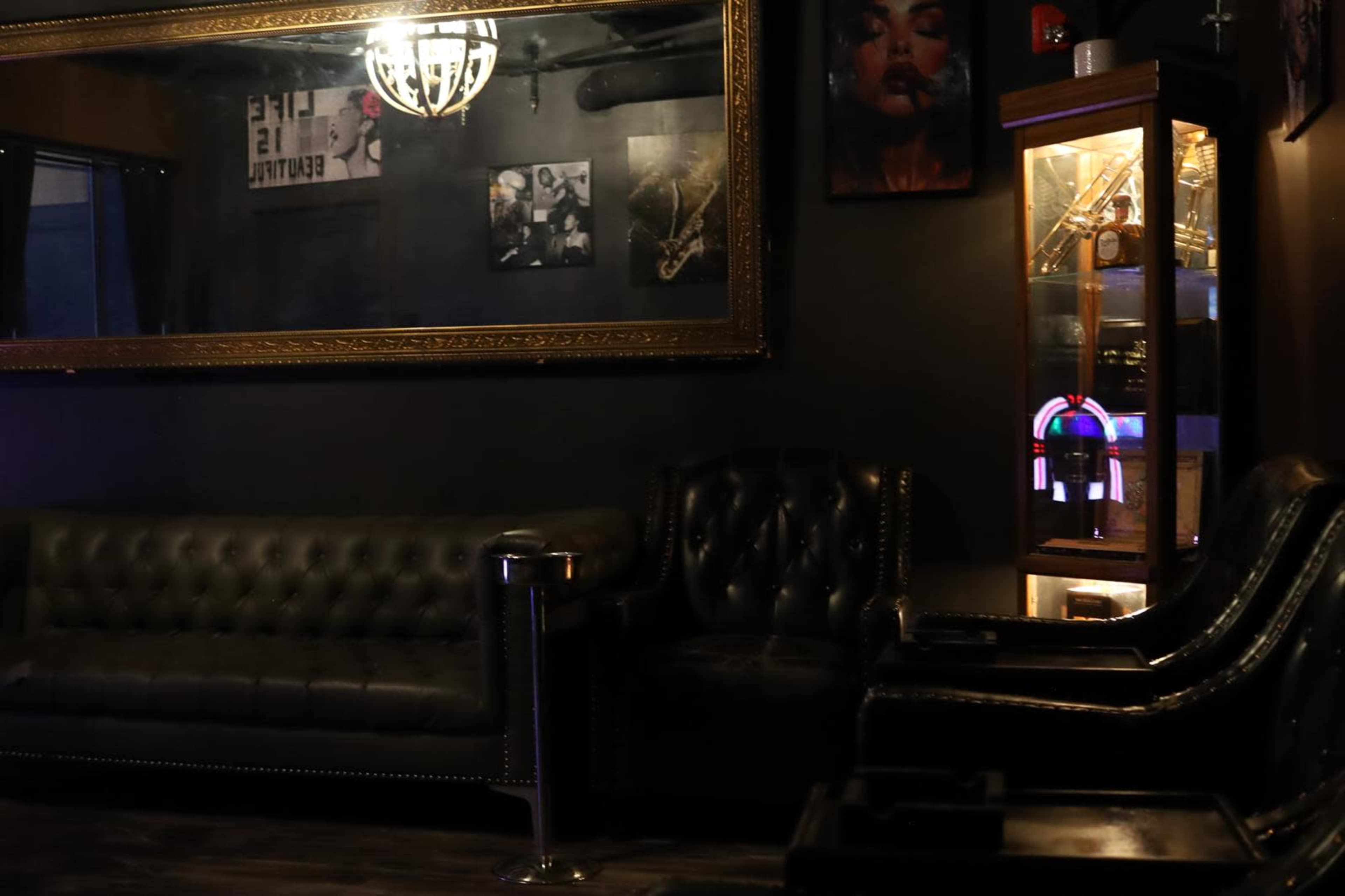 The image depicts a dimly lit lounge area featuring a black leather couch, a pair of armchairs, a decorative cabinet, and wall art.