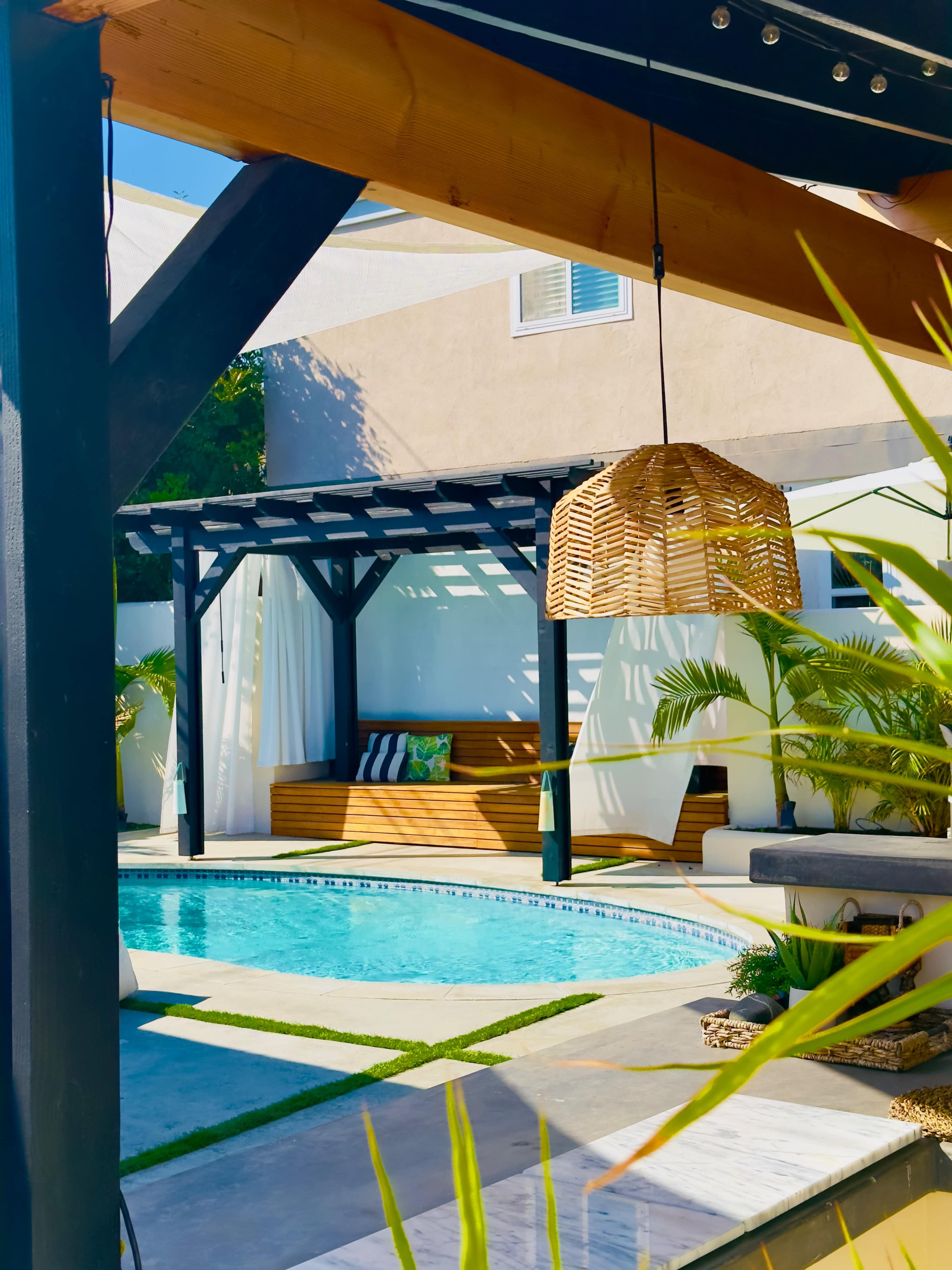 A modern backyard features a swimming pool with a wooden deck, a covered seating area, and lush green plants.