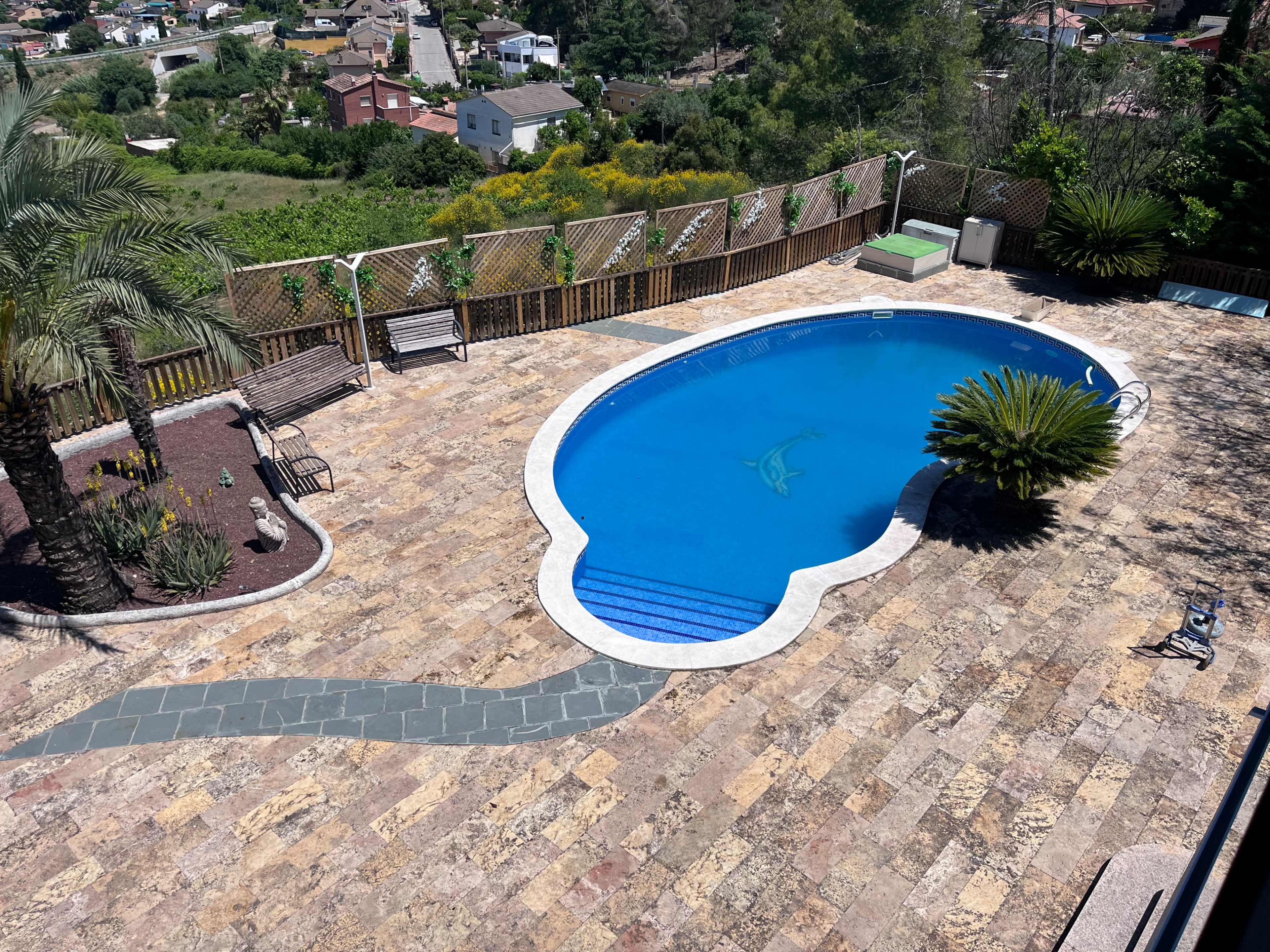 The image shows an aerial view of a swimming pool surrounded by a stone patio and landscaped greenery in a residential setting.