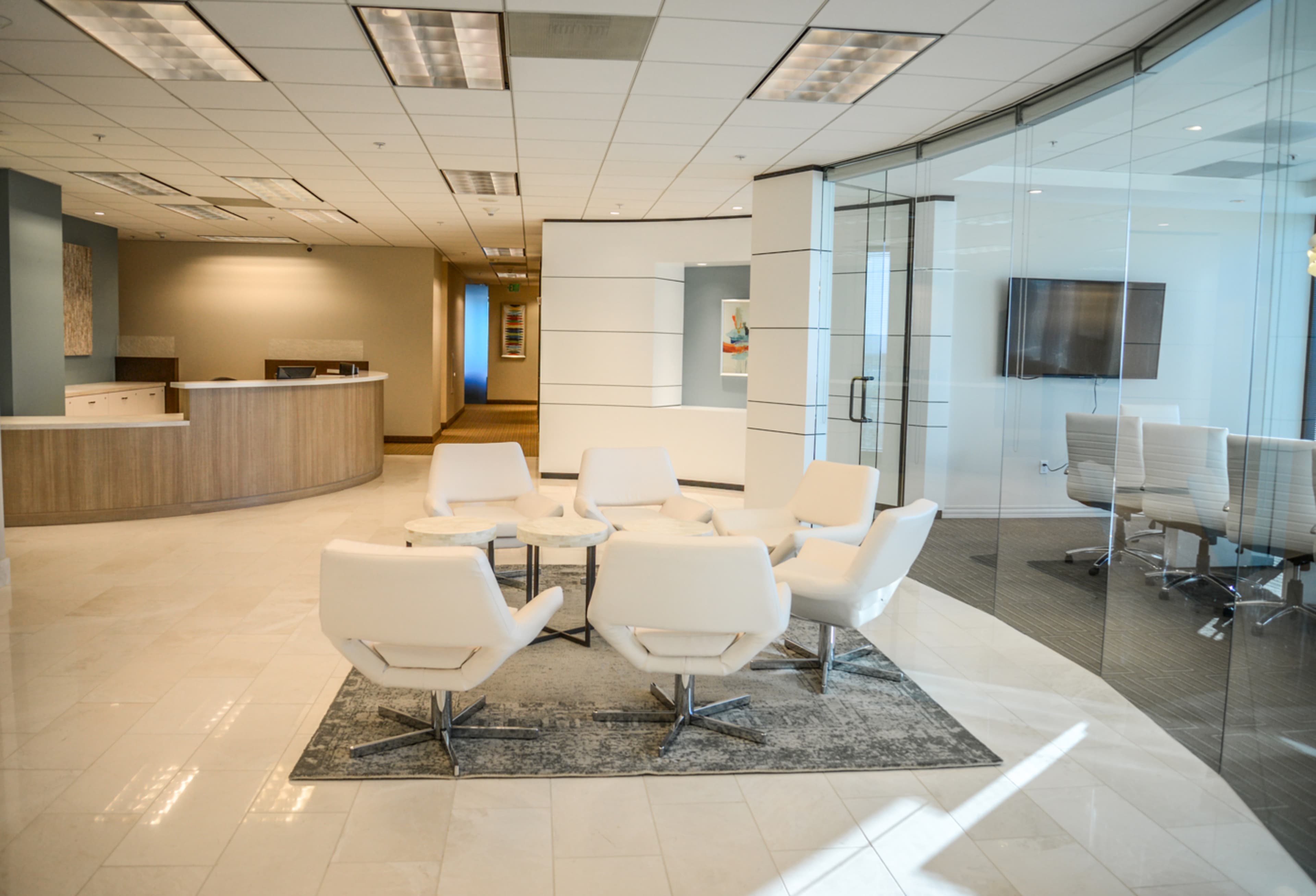A modern office reception area with white chairs arranged around a small table on a rug, and a glass-walled conference room in the background.