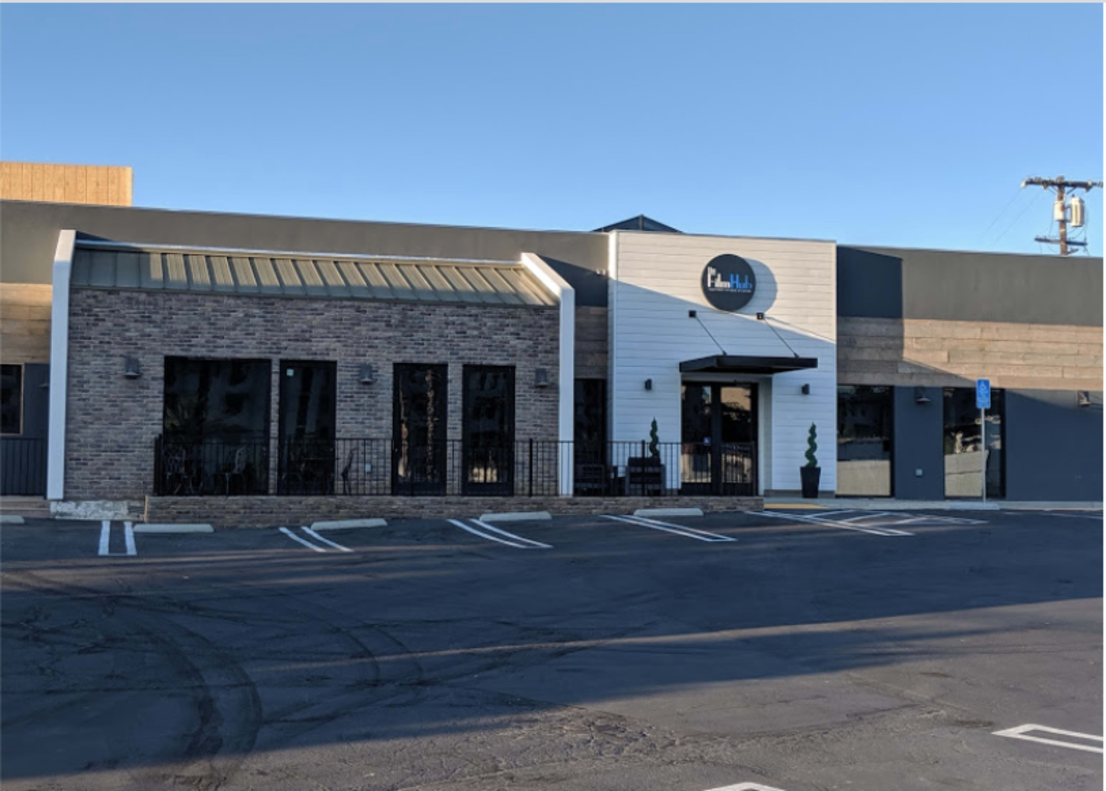 The image shows the exterior of a modern building with a brick facade, a central entrance, and several parking spaces in front.
