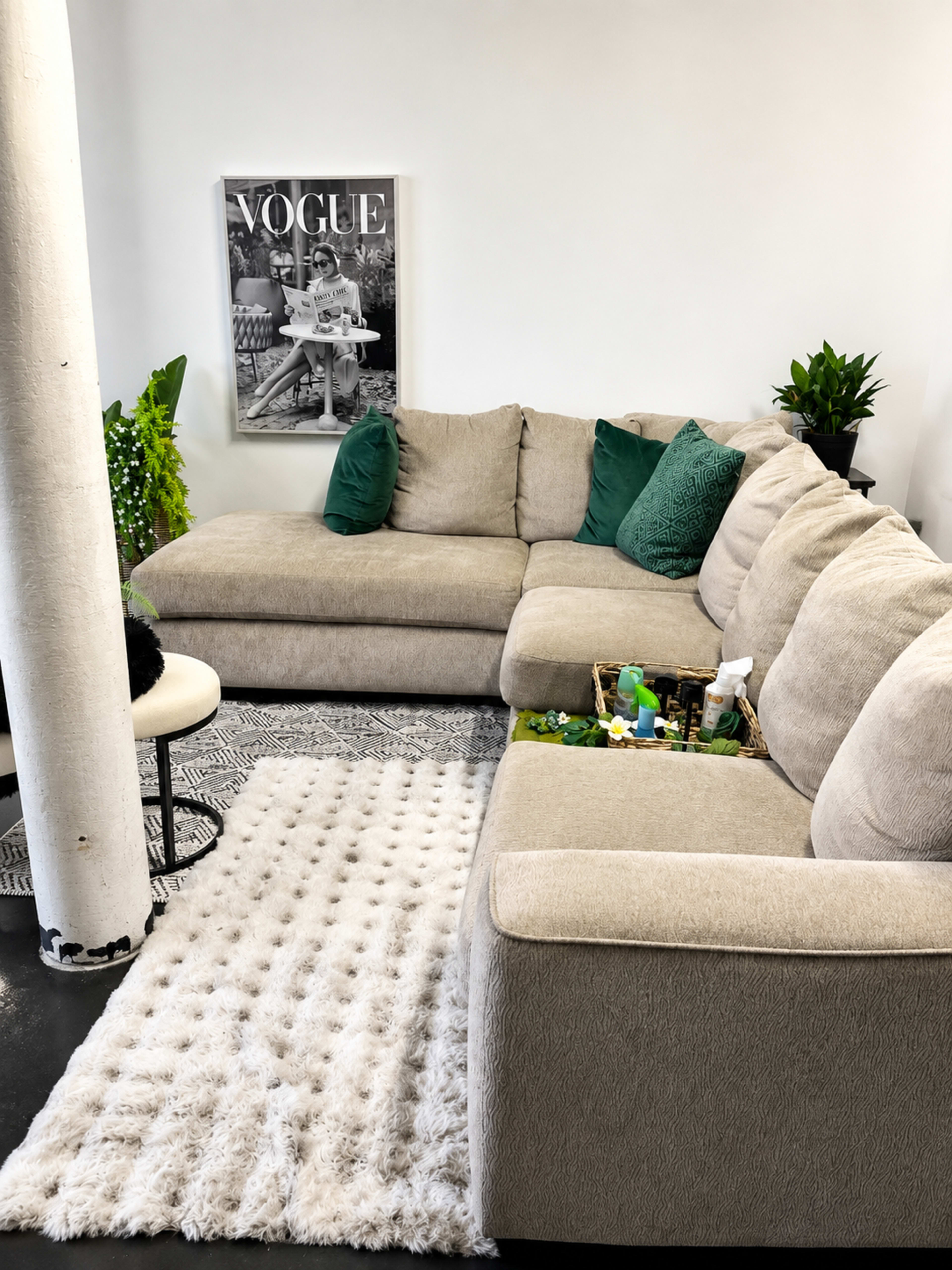 The image shows a modern living room with a large beige sectional sofa, a white shag rug, and a wall-mounted magazine cover featuring "VOGUE" alongside green decorative cushions and indoor plants.
