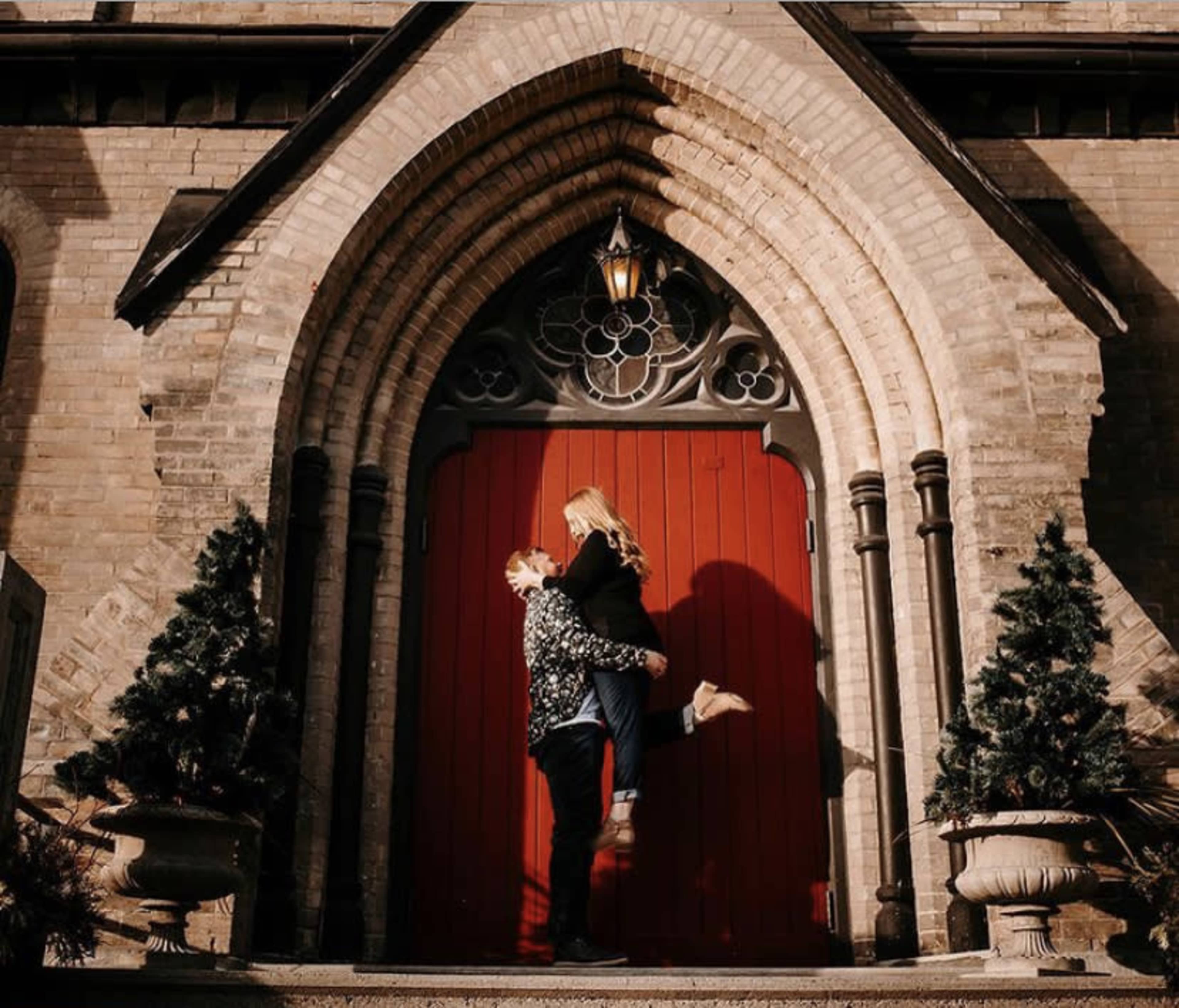 A couple poses in front of a large red door of a stone church, with one partner lifting the other off the ground.