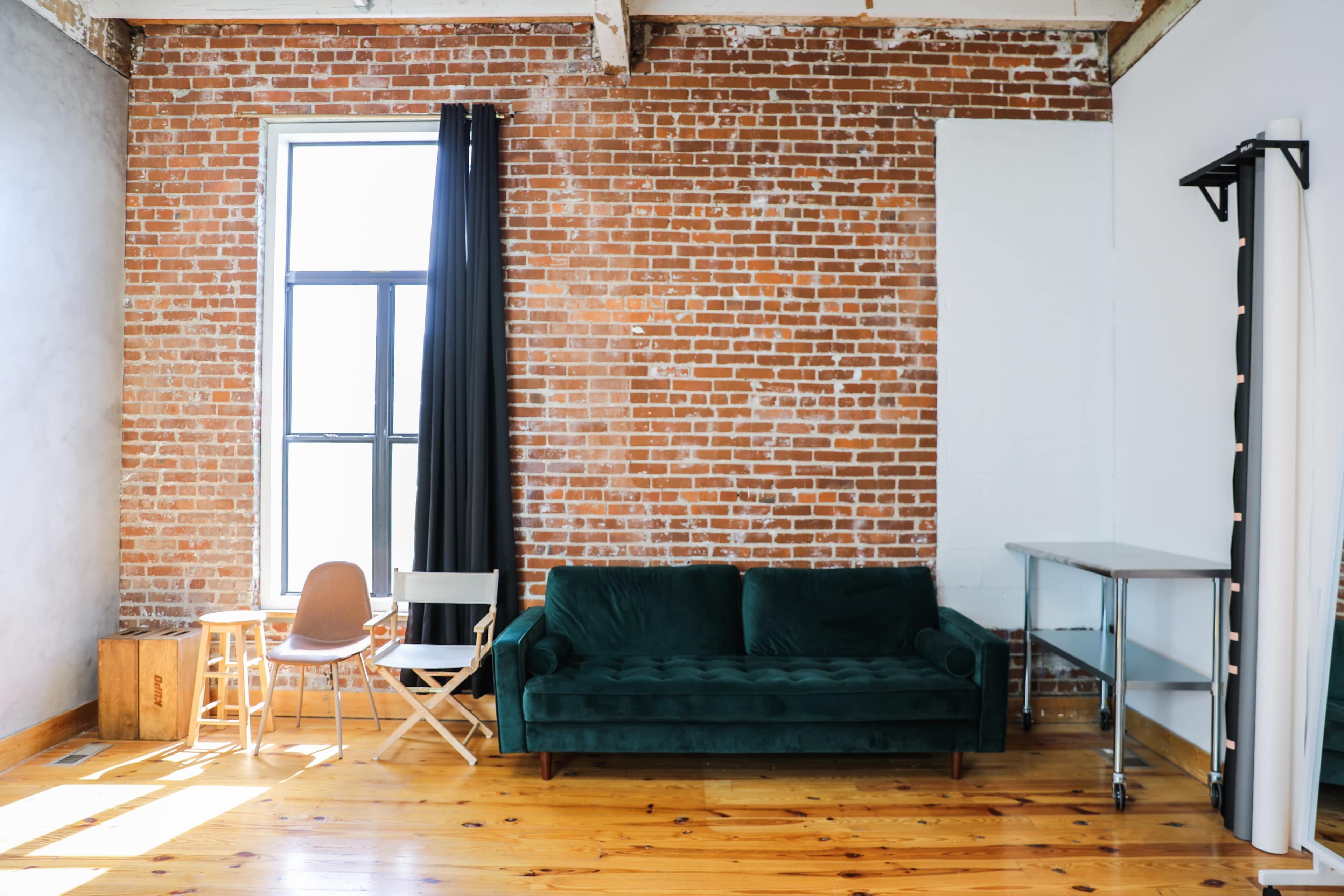 A cozy living space with a green velvet sofa, a wooden chair, a folding chair, and a metallic table against a exposed brick wall.