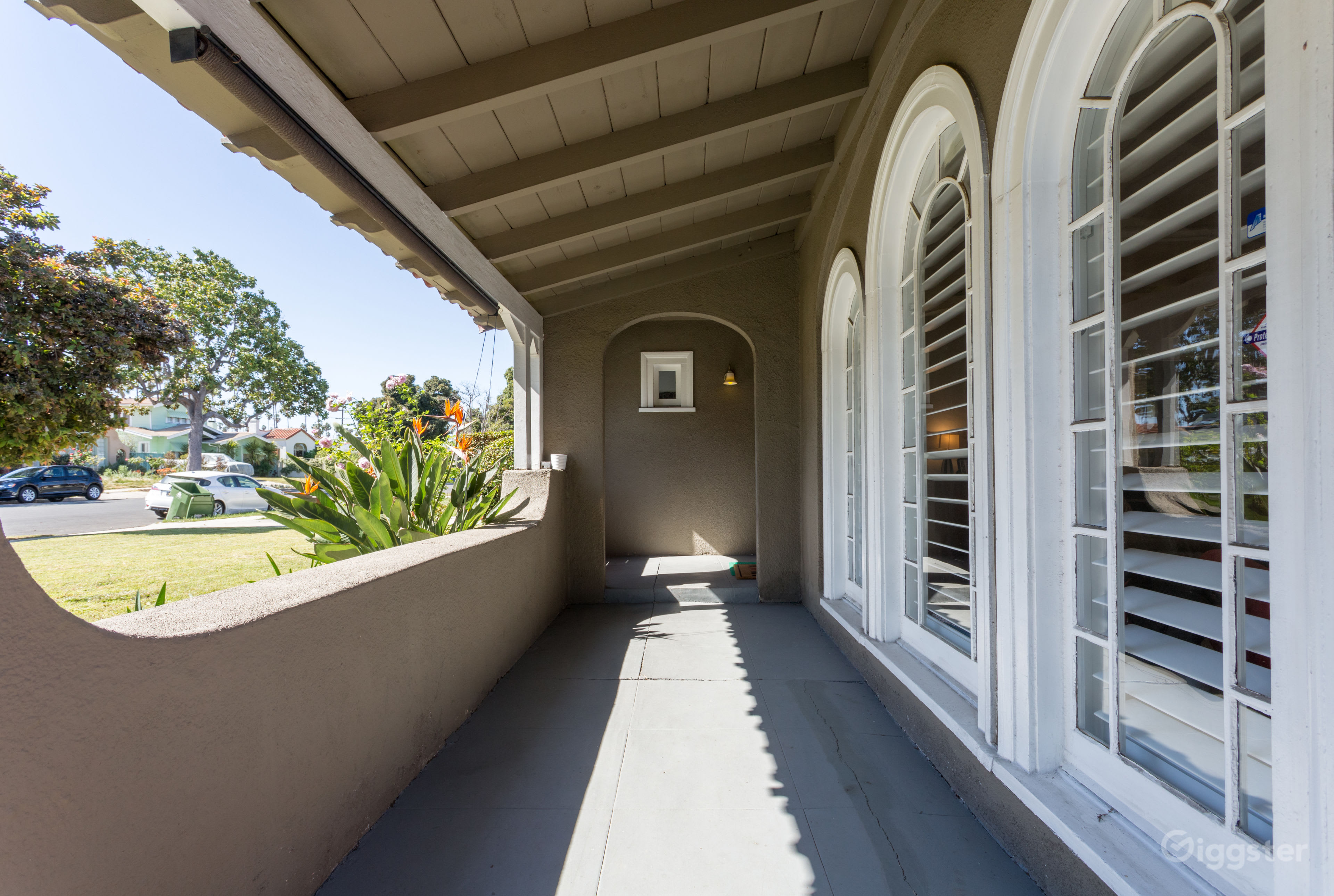 The image shows a covered porch with an arched entrance and large windows, leading to a house and a view of a suburban street.