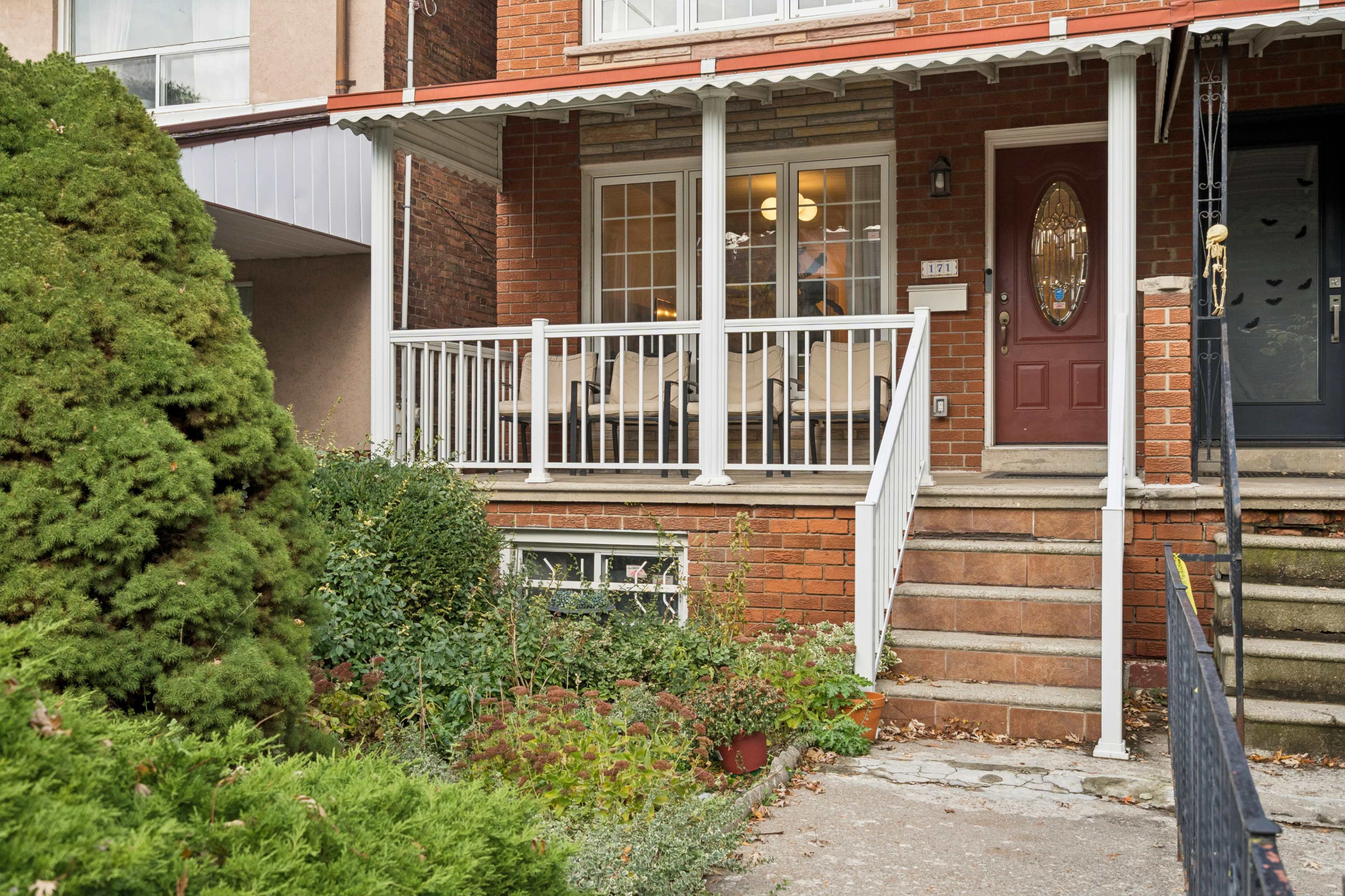 The image shows a brick house with a front porch adorned with white railings and steps leading up to a door, surrounded by greenery.
