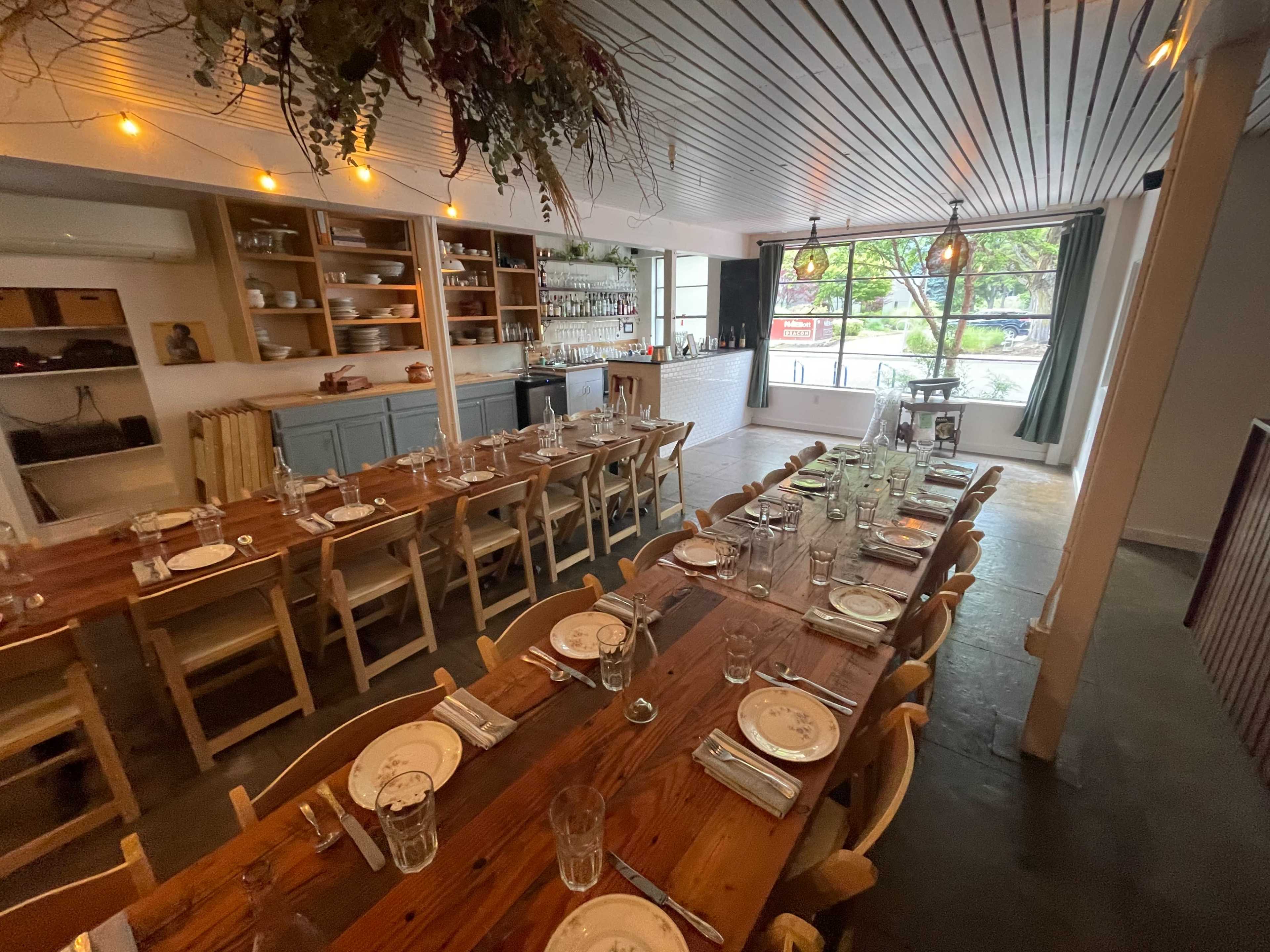The image shows a spacious dining area with wooden tables set for a meal, featuring light fixtures and a large window providing natural light.