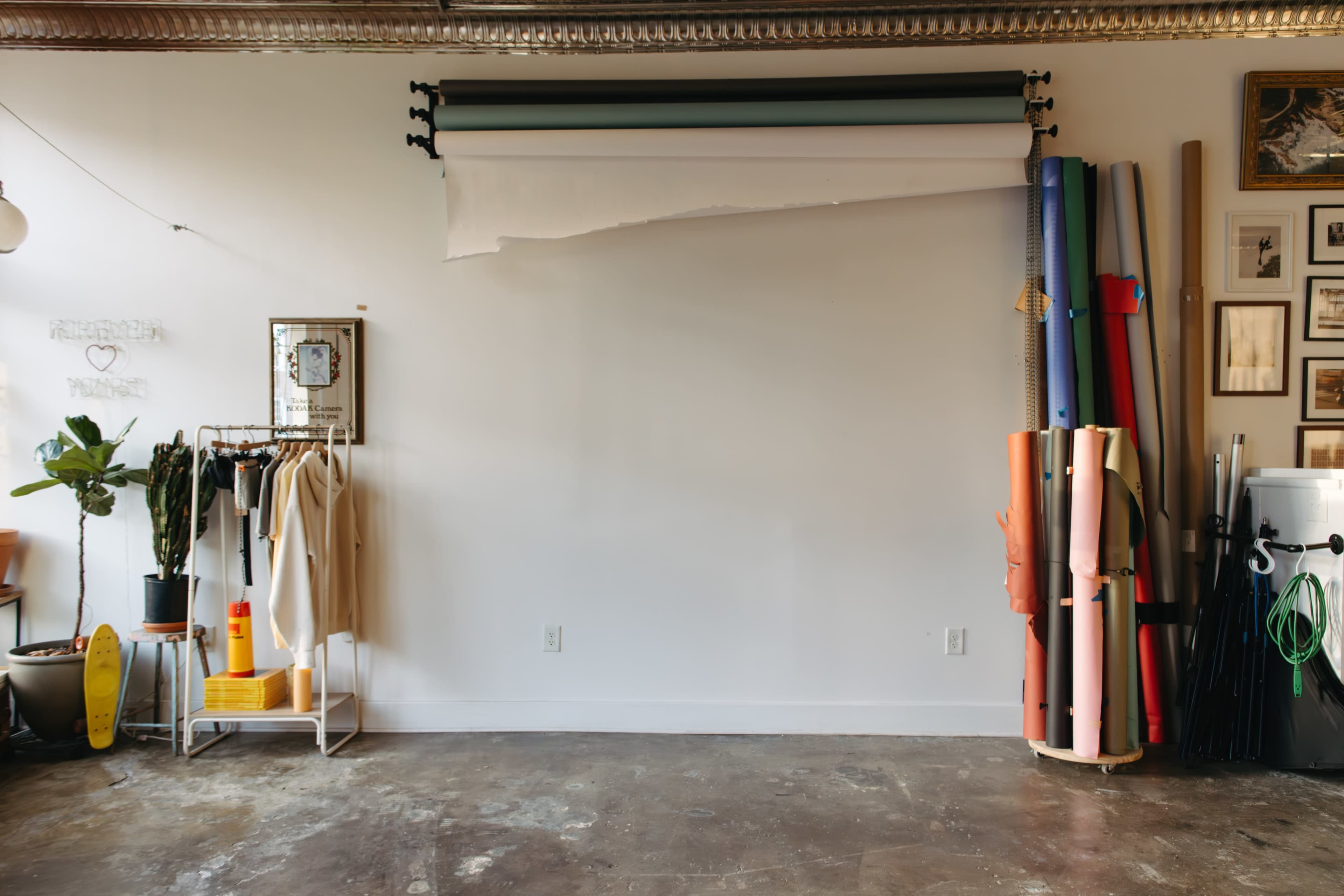The image shows a minimally furnished studio space with a backdrop roll, clothing rack, potted plant, and various colored rolls of material stacked against the wall.