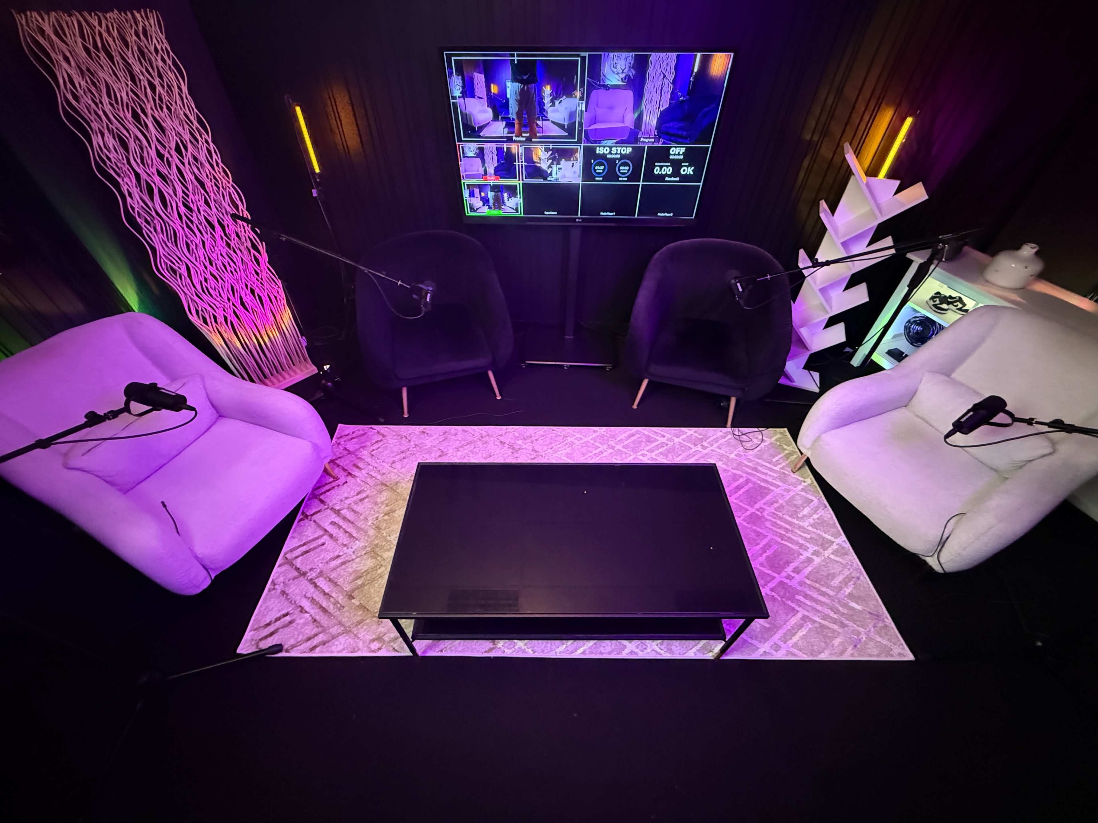 The image shows a cozy interview setup with three chairs, a central coffee table, and a screen displaying video feeds, all surrounded by colorful lighting.