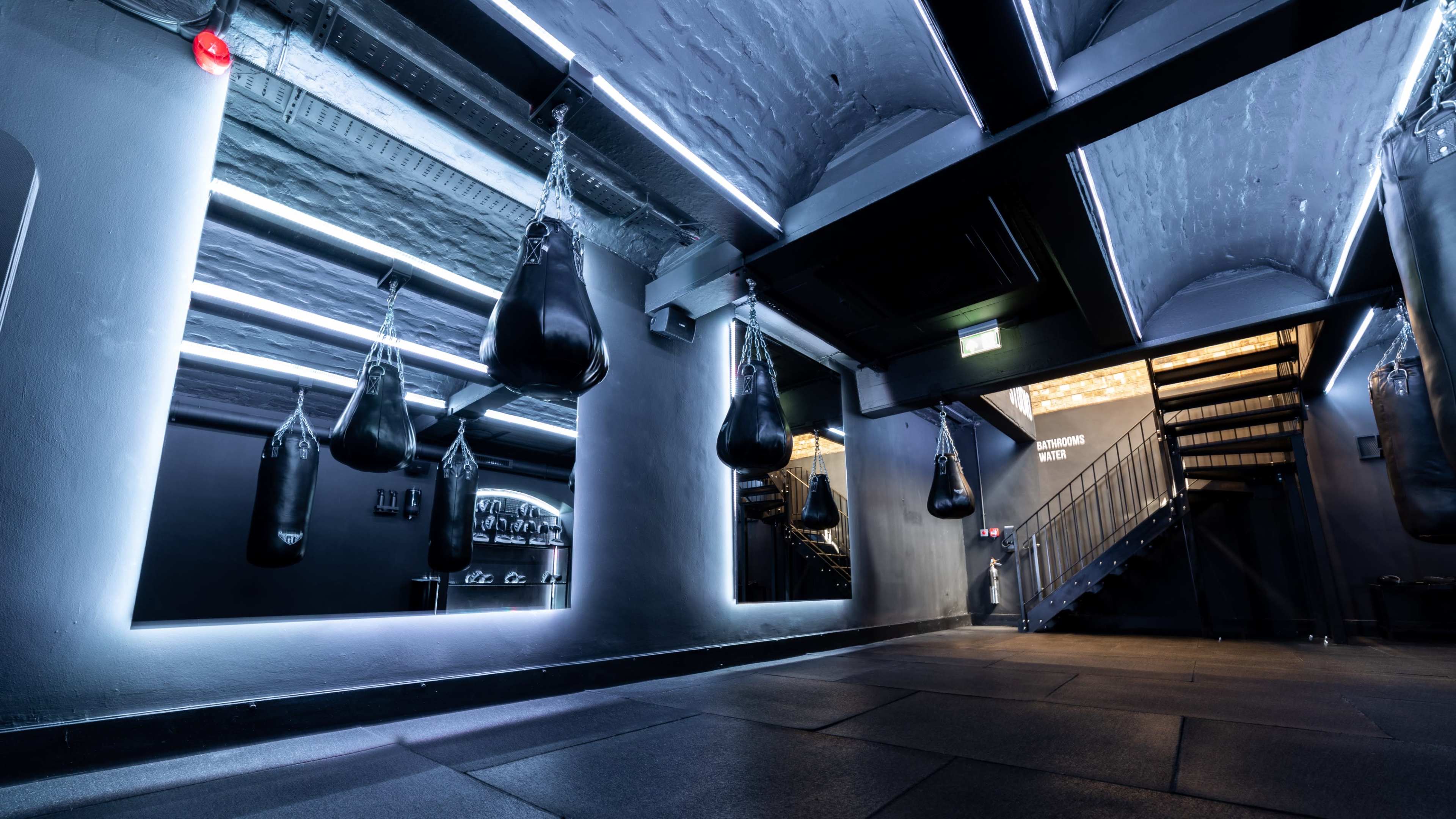 The image shows a dimly lit gym interior featuring several hanging punching bags, mirrored walls, and a staircase.