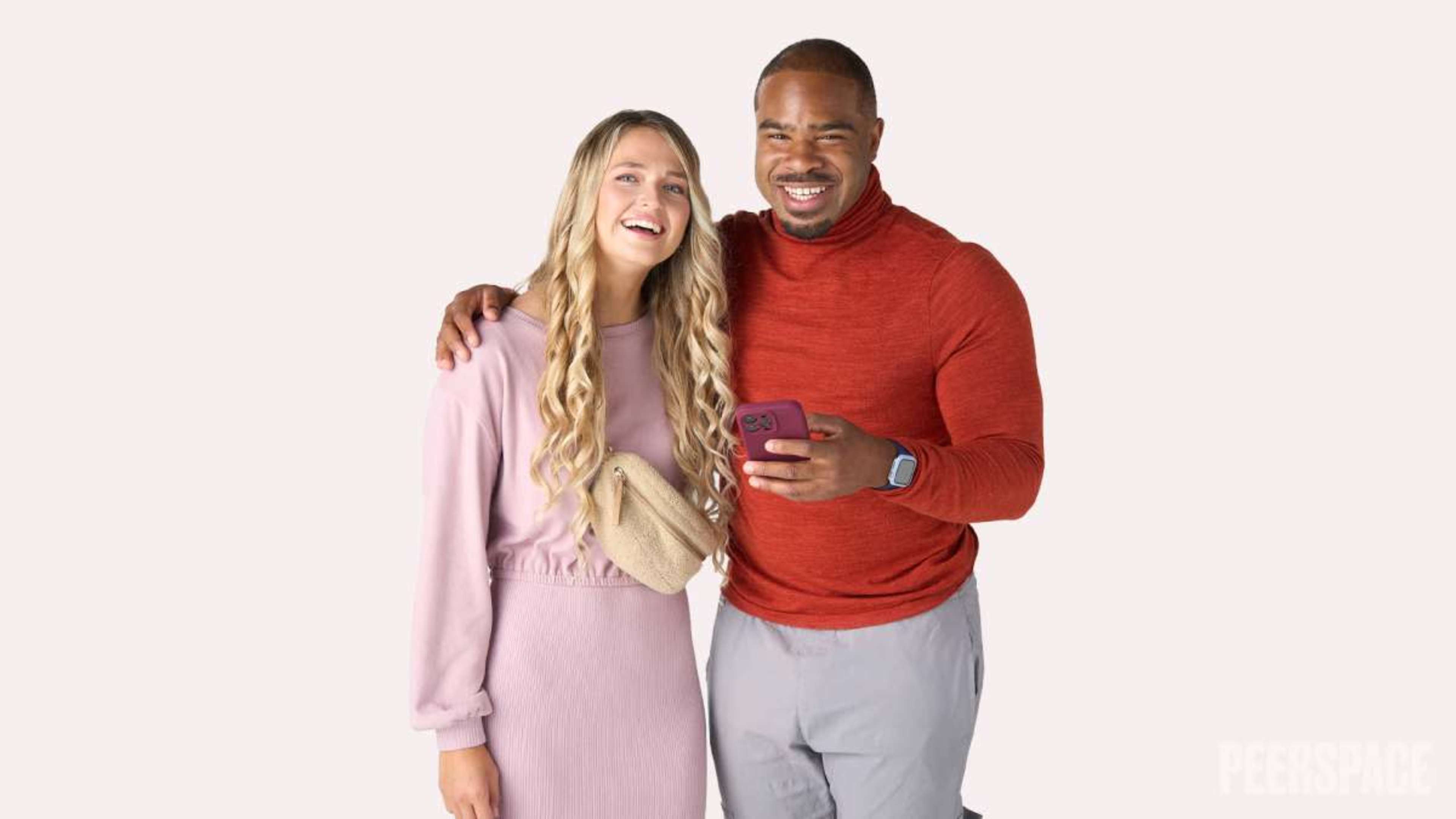 A young woman and a man are standing side by side, smiling, with the woman holding a phone and wearing a light-colored outfit while the man is dressed in a red sweater and gray pants.