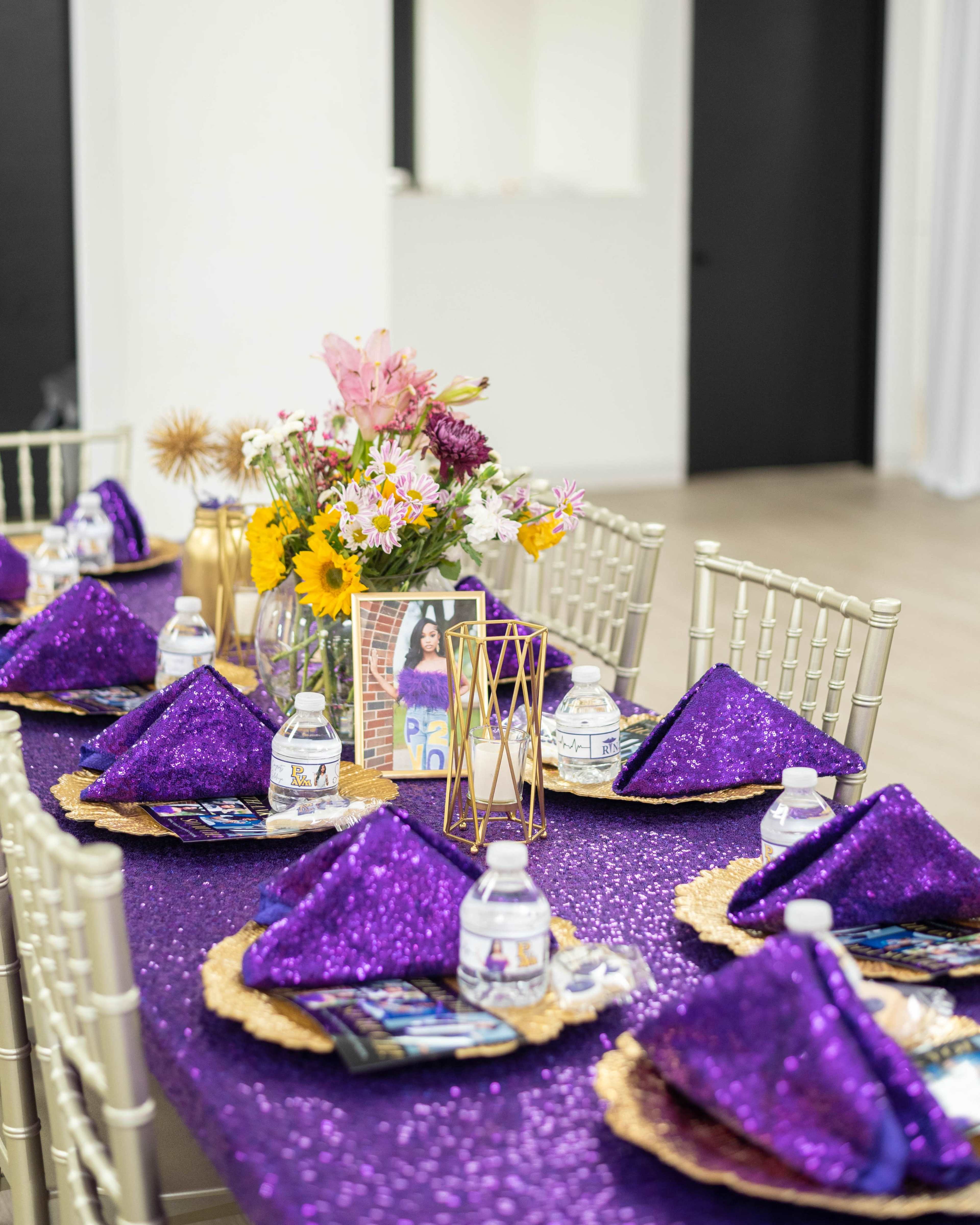 A dining table is set with purple sequin tablecloths, gold chargers, folded napkins, water bottles, and a floral centerpiece.